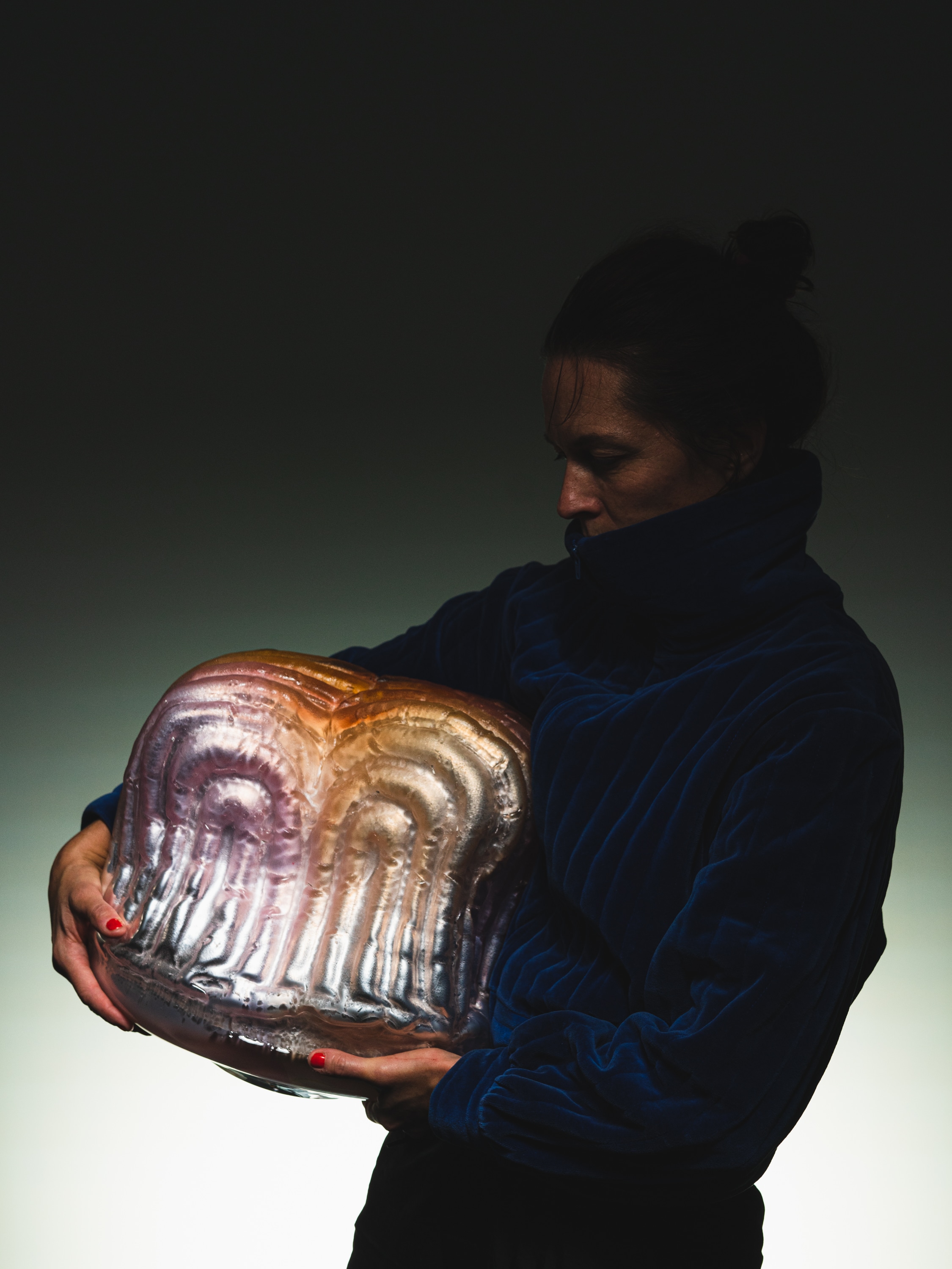 Malin Pierre in a dark blue velvet jacket holds the arched pink, amber and silver glass form against her chest, lit from the side against a dark backdrop.