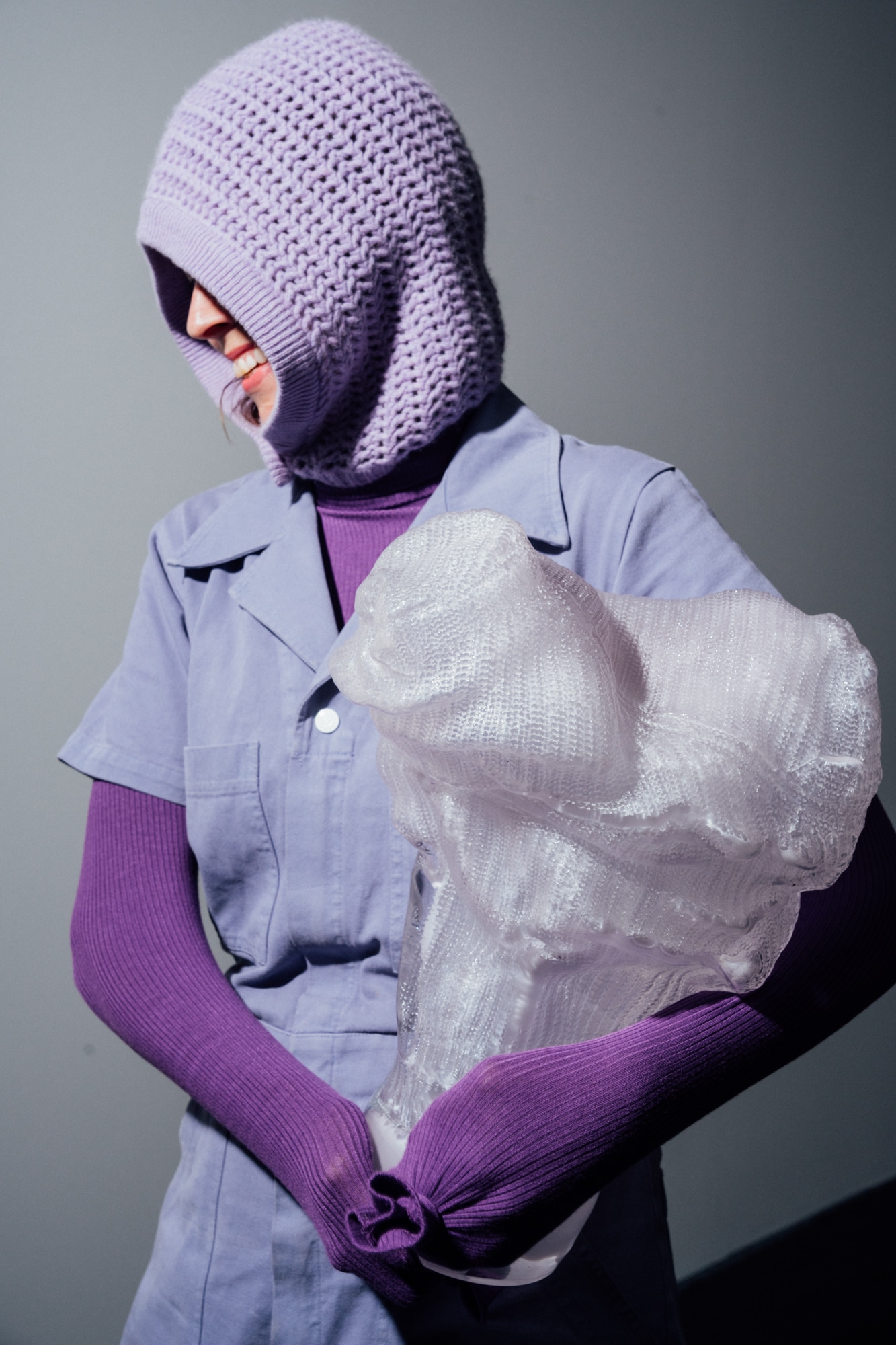 Malin Pierre in a lilac knitted hood and lavender shirt cradles a large clear glass sculpture with knitted-textile imprints, long purple ribbed sleeves knotted beneath it, on a grey backdrop.