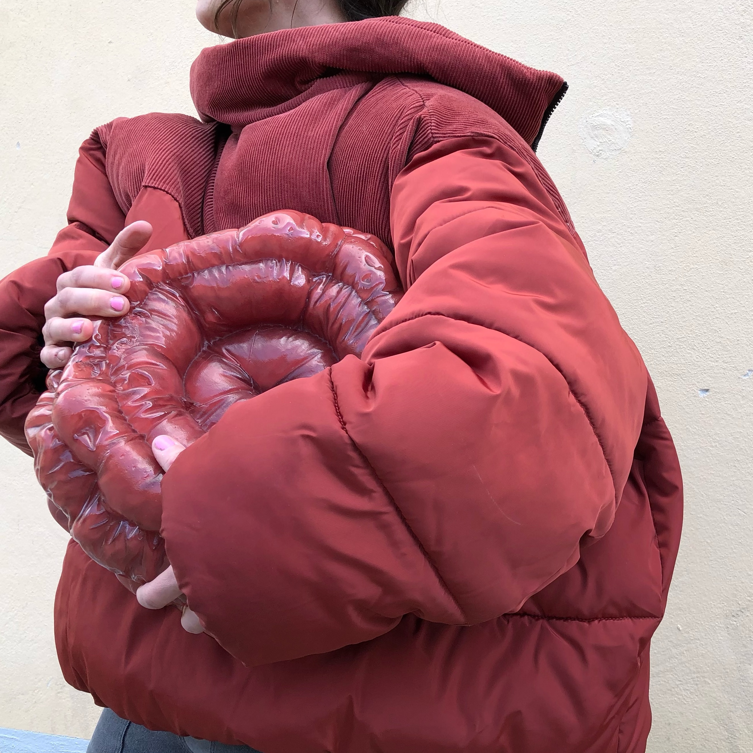 Malin Pierre in a rust-red puffer jacket with a corduroy collar holds a glossy deep red glass form of coiled, quilted rings close to her chest, in front of a pale cream wall.