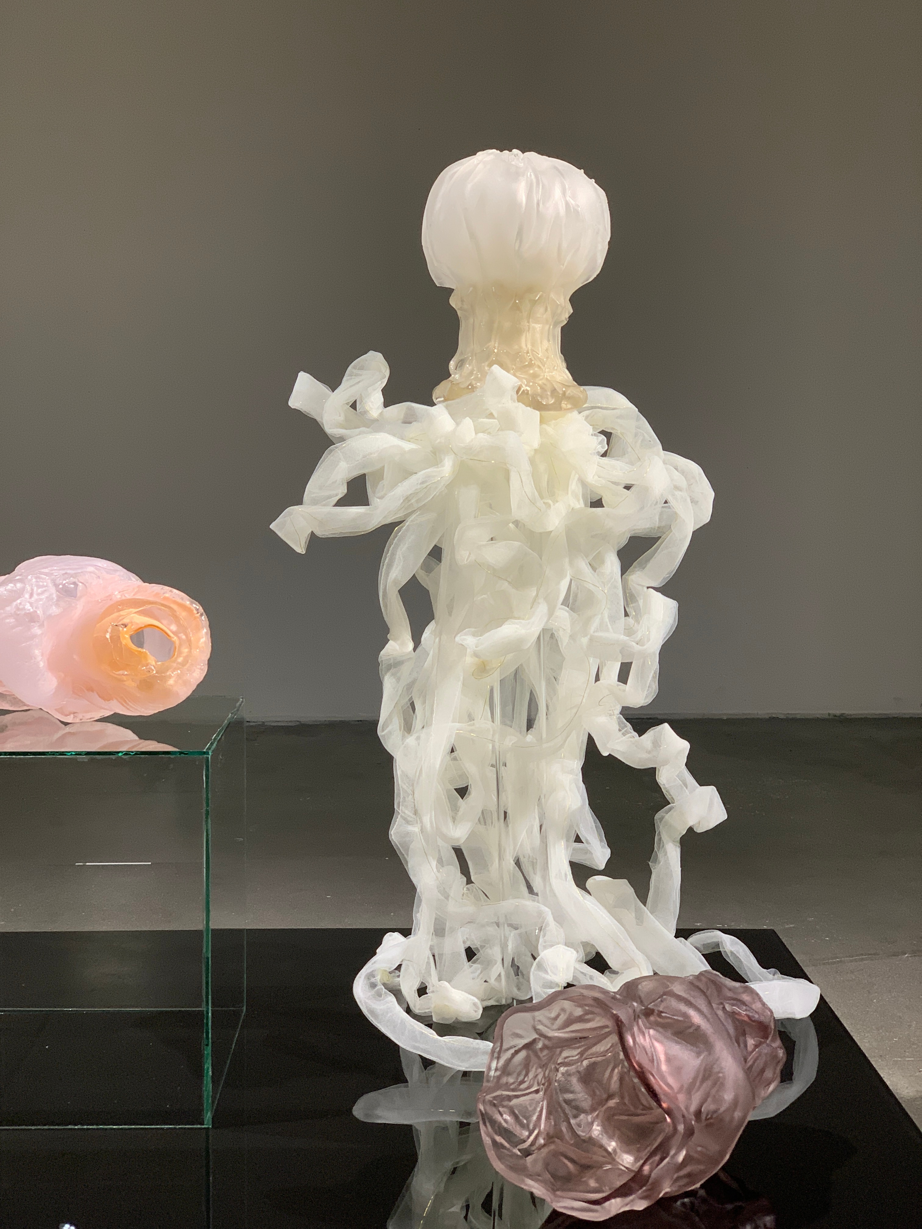 A frosted white glass head on a rippled neck above a cascade of white tulle ribbons, on a black glass platform with a pink and amber coil on a glass cube and a mauve form at its foot.