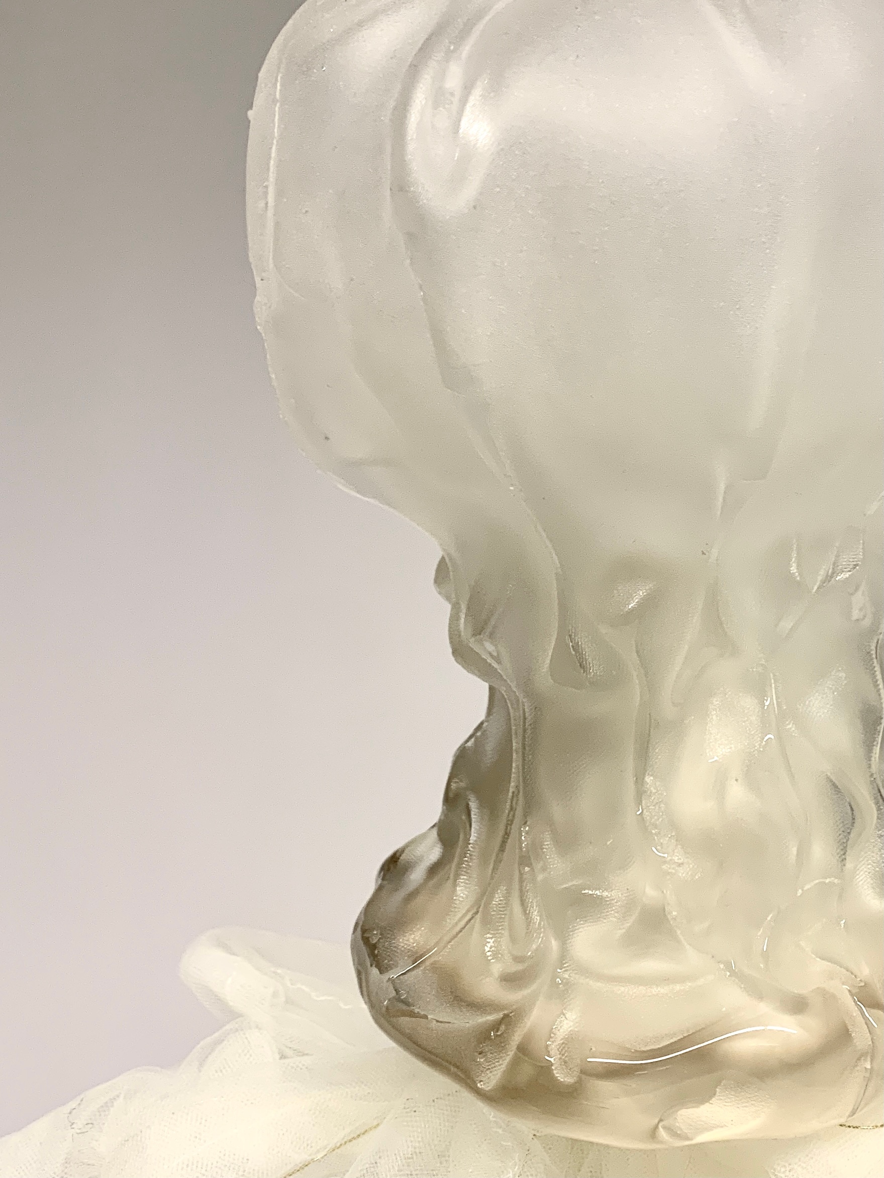 Close-up of a frosted white glass form with a rounded head and a glossy, rippled neck in clear and pale amber glass, rising from white tulle against a grey wall.