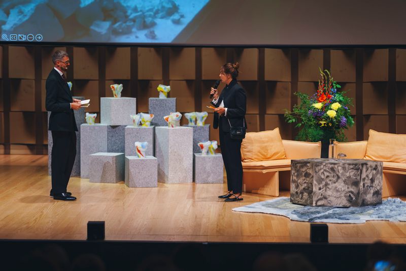 The award ceremony on a stage of light wooden panels: a man with papers and a woman with a microphone stand among grey stone plinths holding small glass award sculptures, beside peach sofas and a large bouquet.
