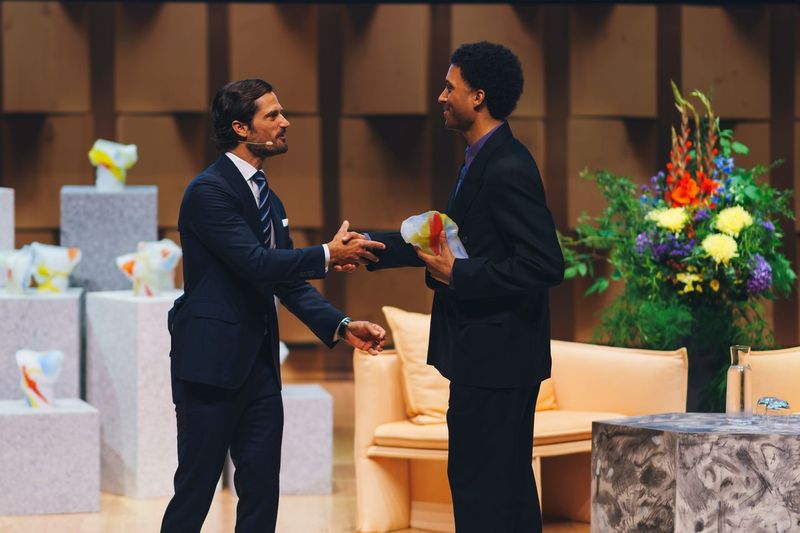 Two men in dark suits shake hands on stage; one holds a white glass award sculpture streaked with red and yellow, and more of the awards stand on grey stone plinths behind them.