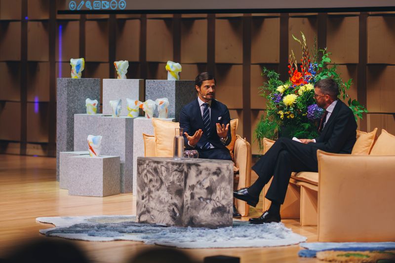 Two men in suits talk on peach sofas on stage, with the glass award sculptures on stacked grey stone plinths behind them and a tall bouquet of summer flowers.