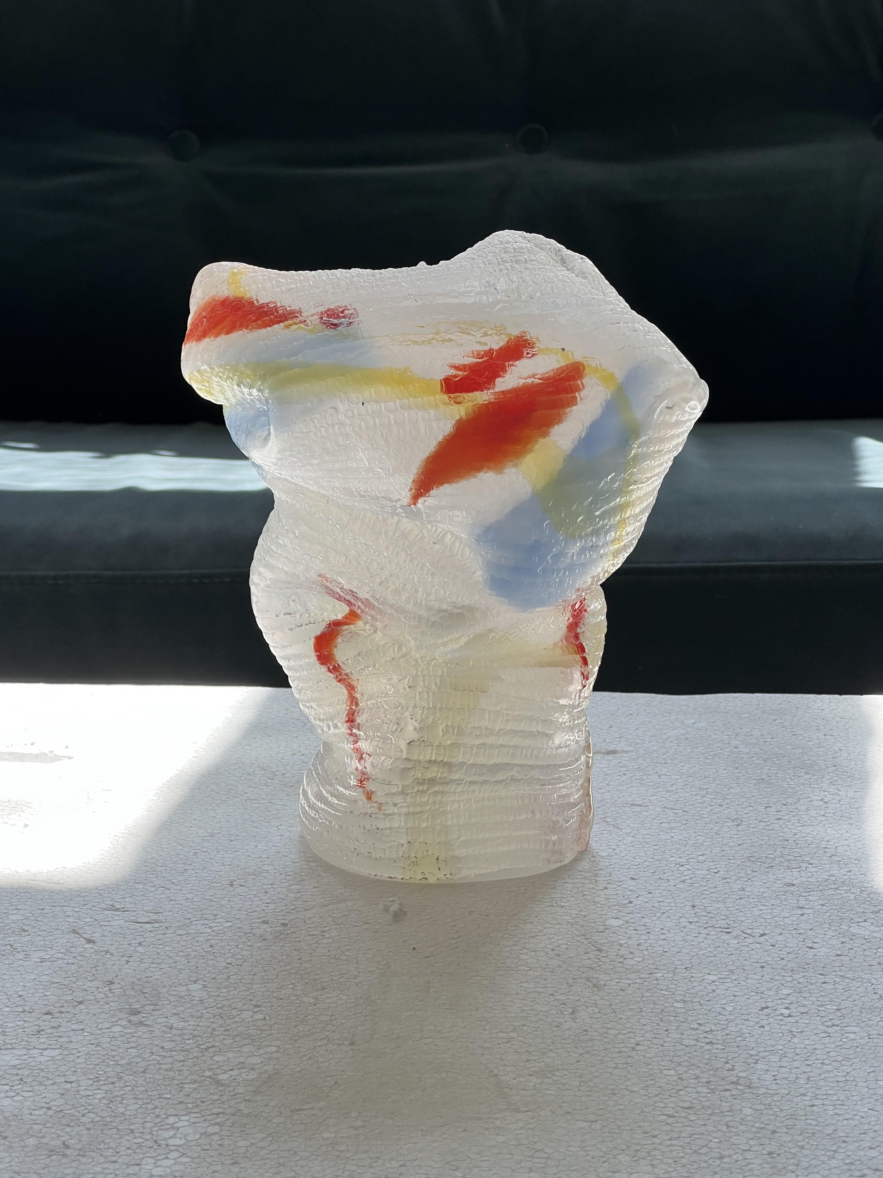 One of the Utmärkt Svensk Form award sculptures: a crumpled, knit-textured form of clear glass streaked with red, yellow and pale blue, standing in sunlight on a white table in front of a dark green sofa.