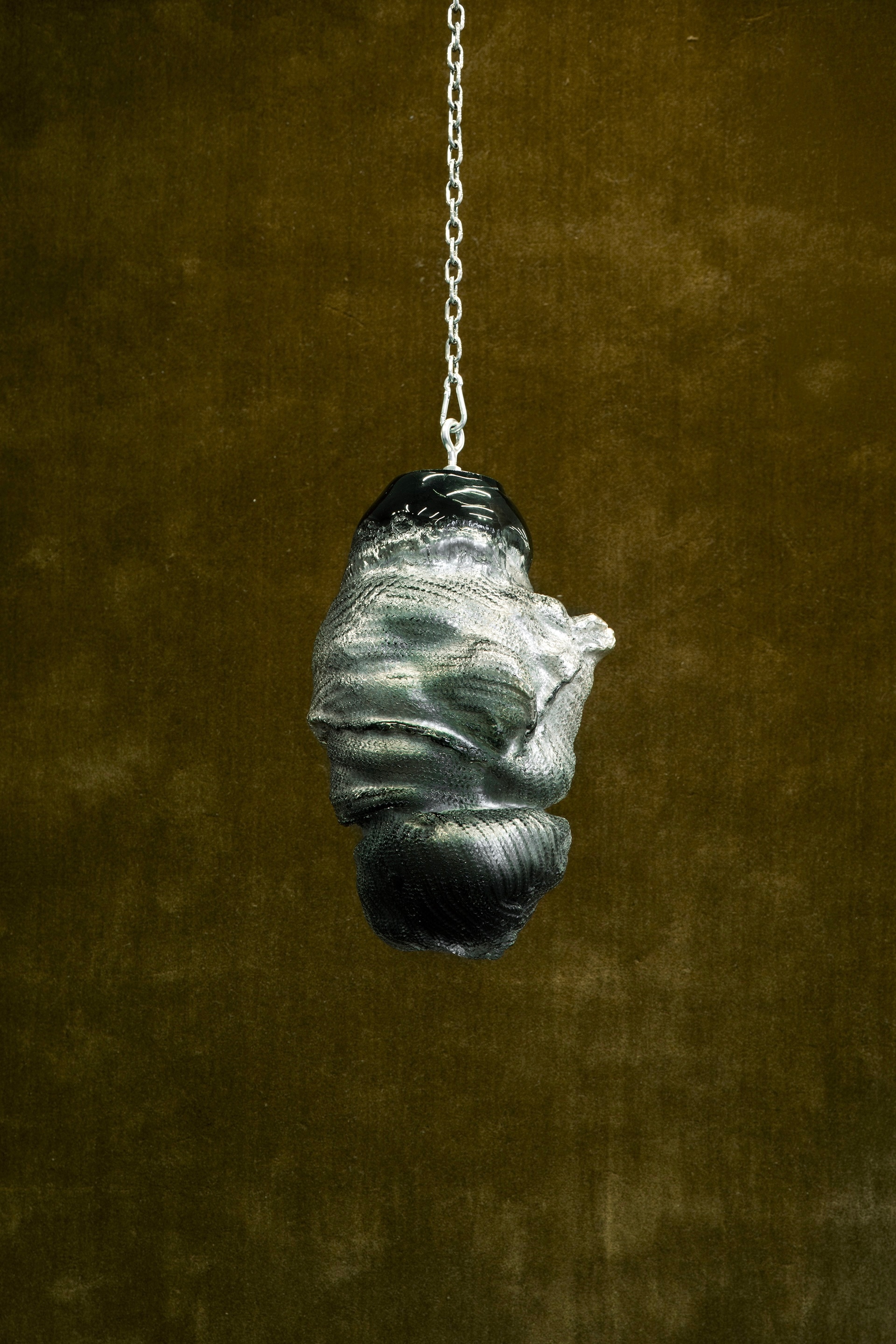 A glass sculpture hanging from a steel chain: a crumpled, bundled form with knitted-stitch imprints, mirror-silver shading to black at the top and bottom, against an olive green velvet backdrop.