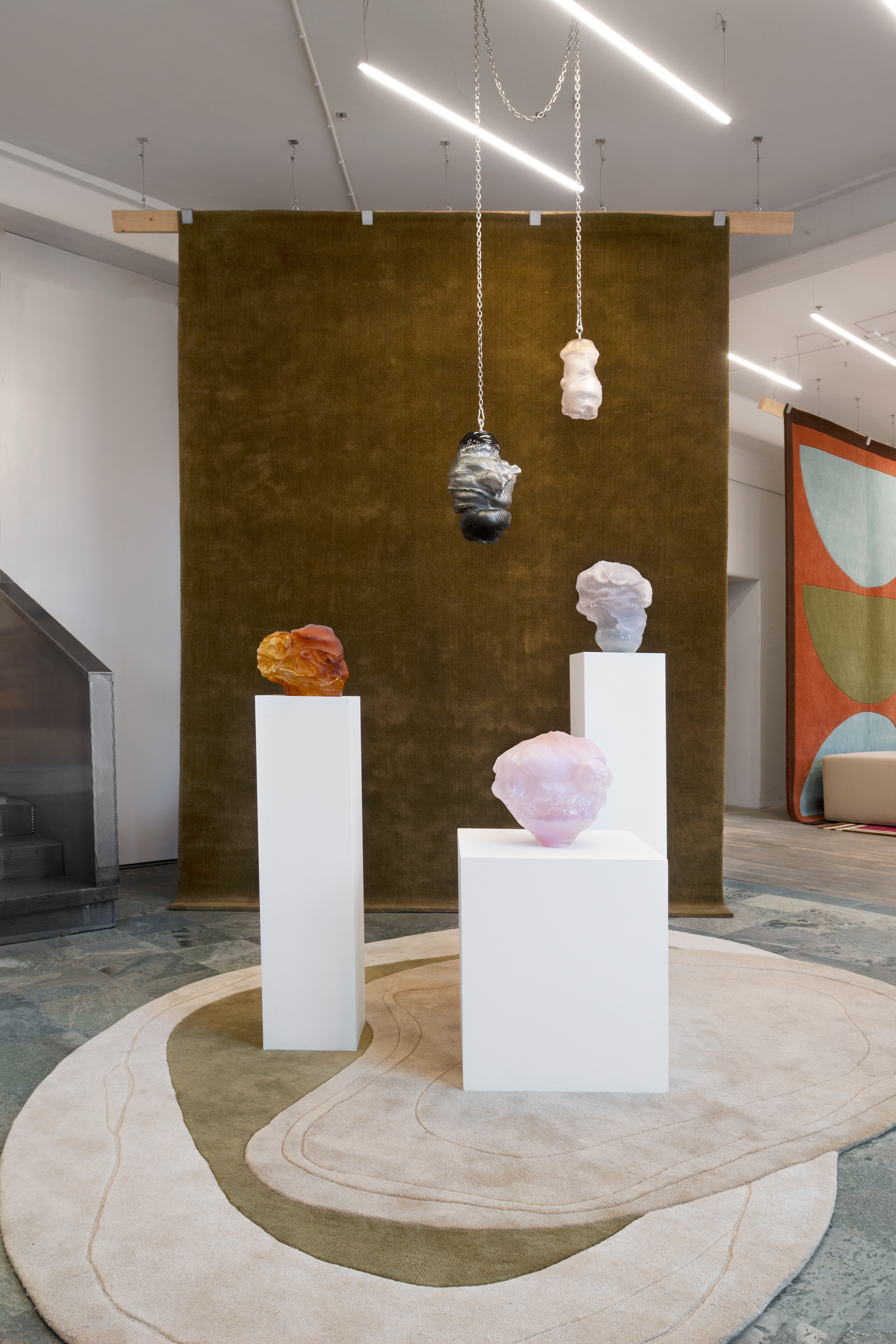 Exhibition view: five glass sculptures in front of a moss-green rug hung as a backdrop, an amber and a lilac form on tall white plinths, a pink one on a low plinth, and a black-and-silver and a pale pink form hanging from steel chains, above a cream rug with green contours.