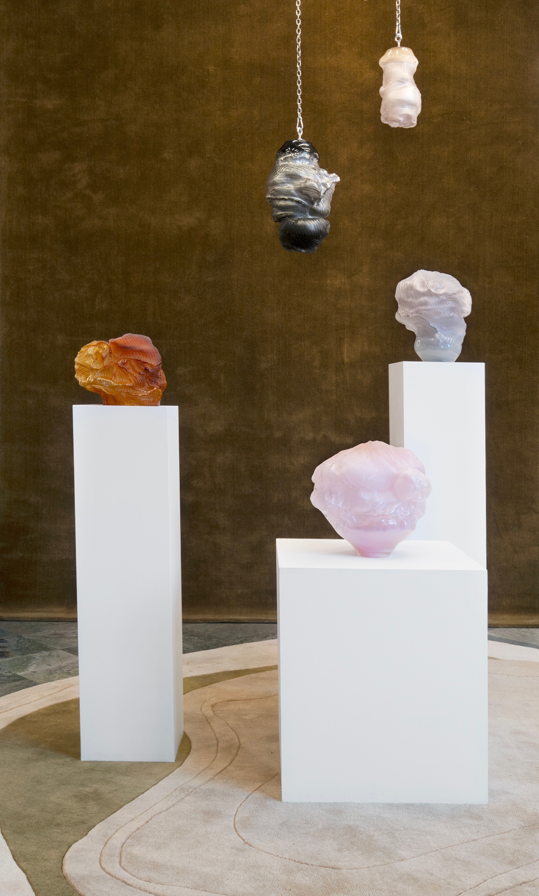 Closer view of the same installation: the amber, lilac and pink glass forms on their white plinths and the black-and-silver and pale pink forms hanging on chains, against the moss-green backdrop.