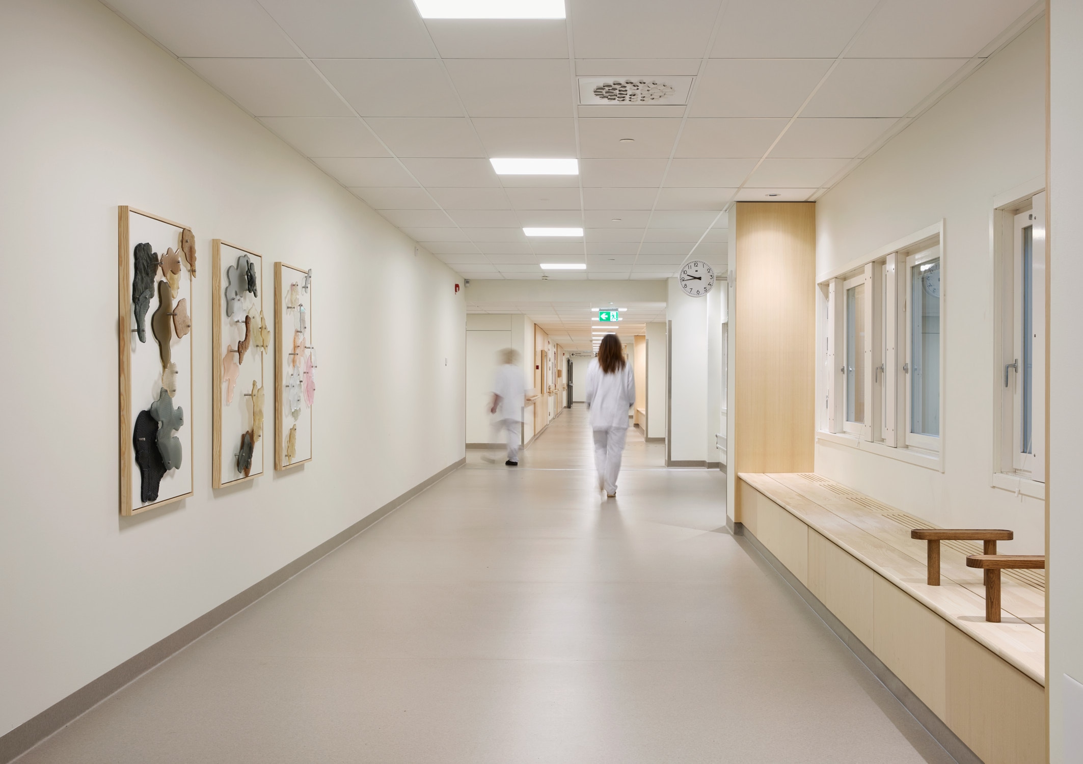 A hospital corridor with three tall light wooden frames on the left wall, each holding clusters of flat glass shapes in black, grey, pink, peach and cream; two staff in white walk away down the corridor.