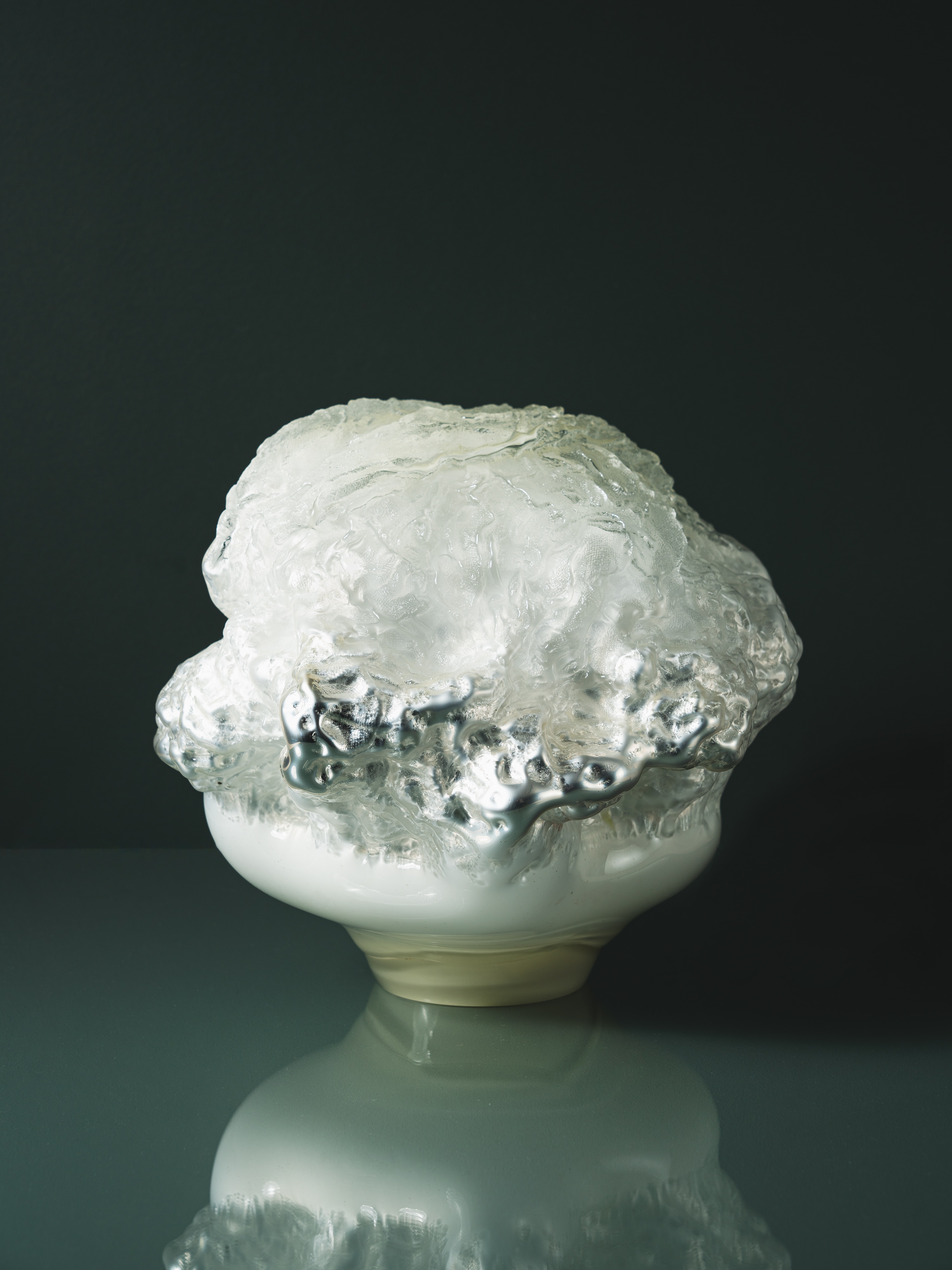 A cloud-like glass form, its frosted, crumpled top edged with mirror-silver drips, on a smooth milky white base, against a dark green-grey wall.