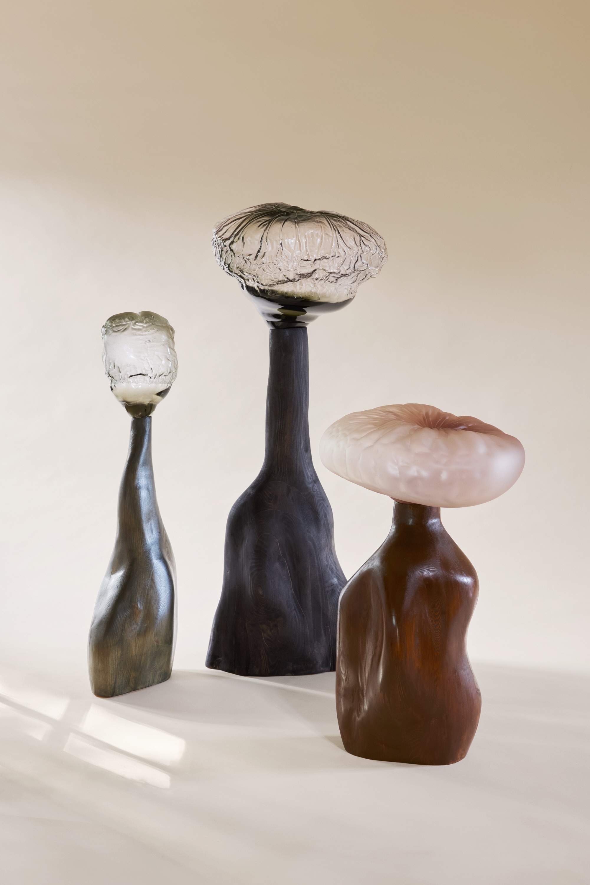 Three sculptures on a cream backdrop: two crumpled smoky-grey glass tops and a frosted blush-pink cushion cap on turned grey-green, charcoal and red-brown wooden stems, in window light.