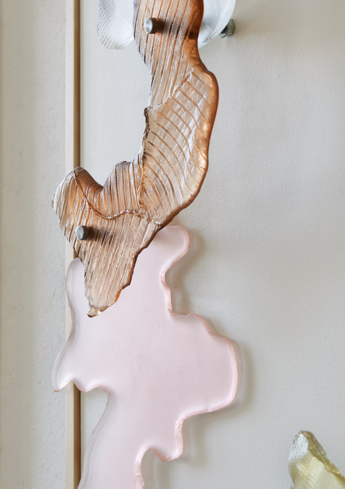 Close-up of a copper-brown glass shape with fine ridged lines, held on steel pins above a frosted pale pink shape, against pale canvas beside a light wooden frame edge.