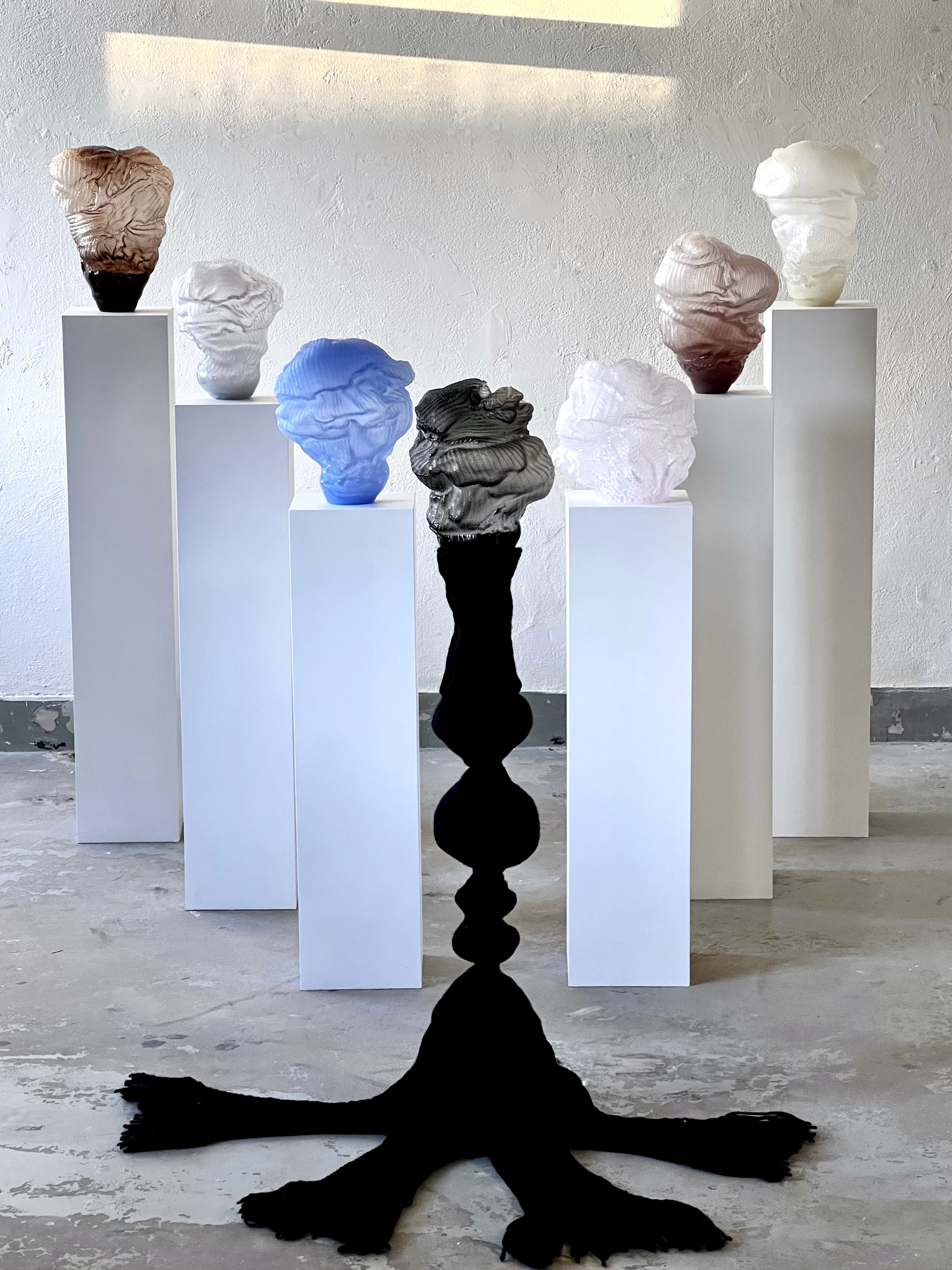 Six glass forms with knitted-stitch textures in smoky brown, white, blue, clear, mauve and ivory on white plinths, behind a dark grey one on a black knitted column with fringed feet on concrete.