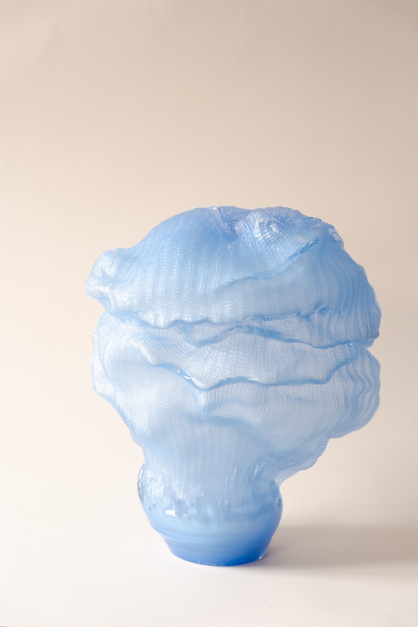 The sculpture Britt-Marie: a frosted cornflower-blue glass form with a knitted-stitch surface in wavy, layered folds, narrowing to a small glossy foot, on a pale peach backdrop.