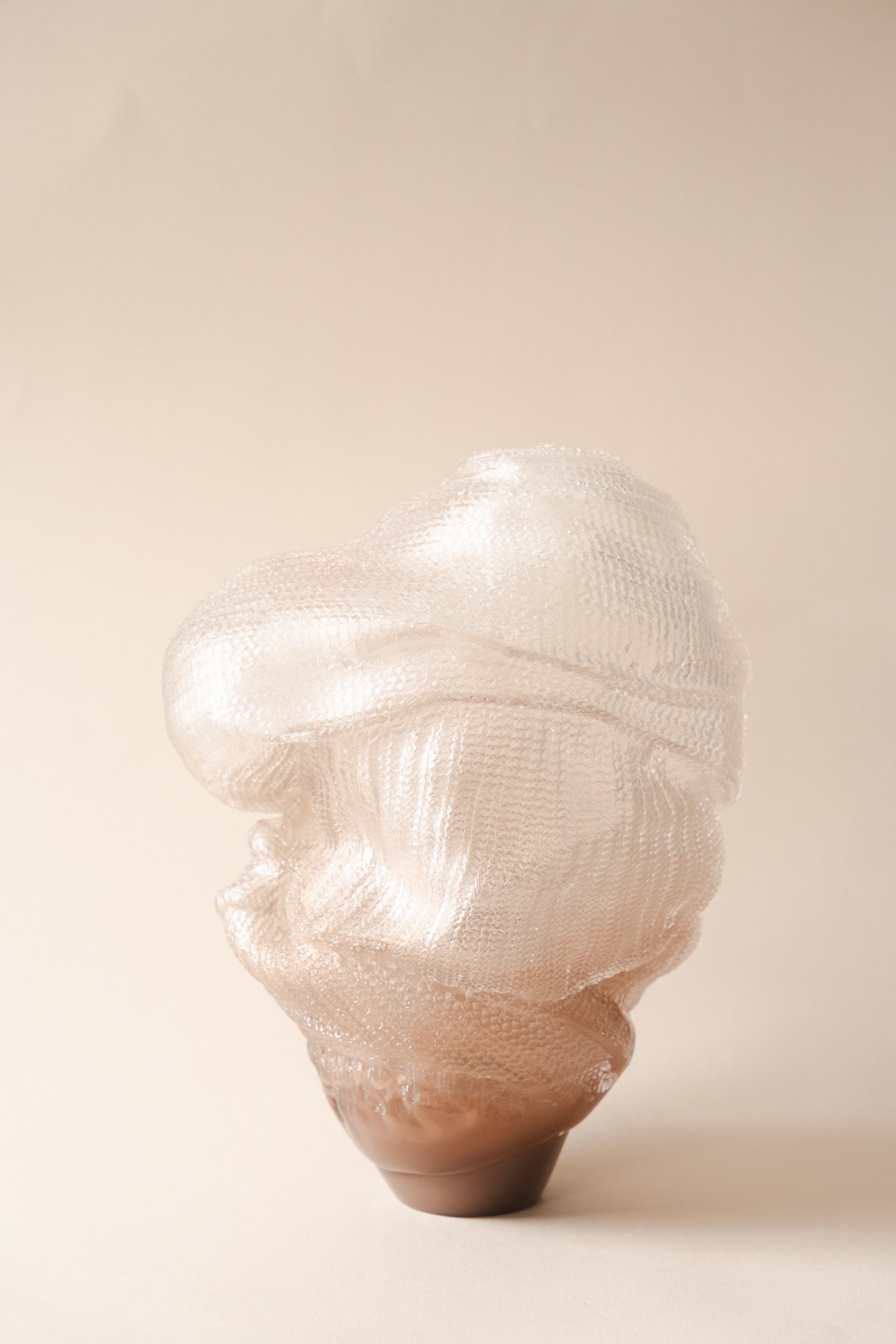 The sculpture Maggan: a near-clear glass form with a knitted-stitch surface folded like draped cloth, shading to a smooth taupe-brown foot, on a soft peach backdrop.