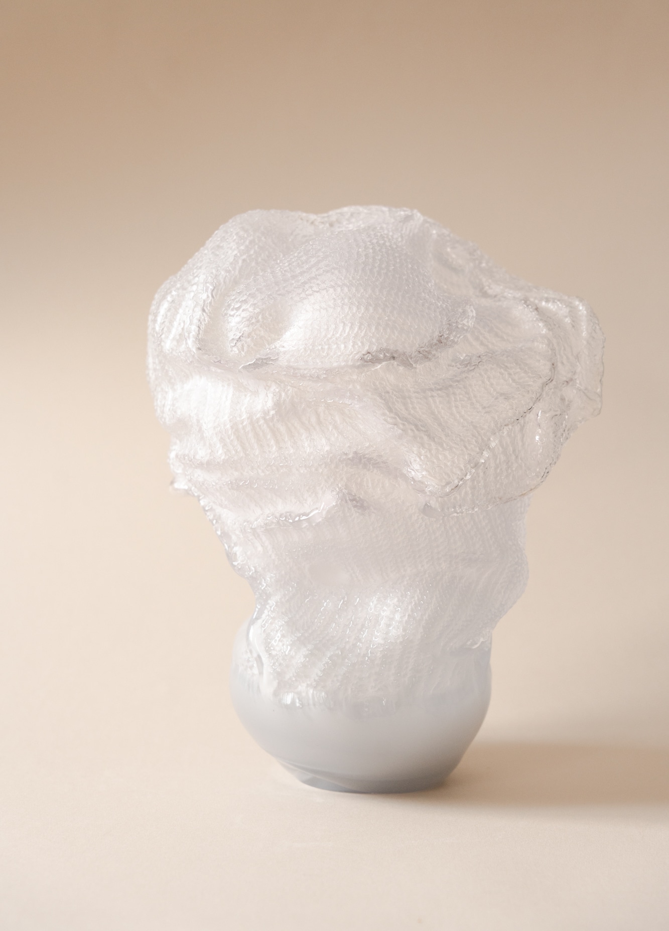 The sculpture Ulla: a clear glass form with a knitted-stitch surface, gathered and folded like loose fabric above a rounded milky grey base, against a peach background.