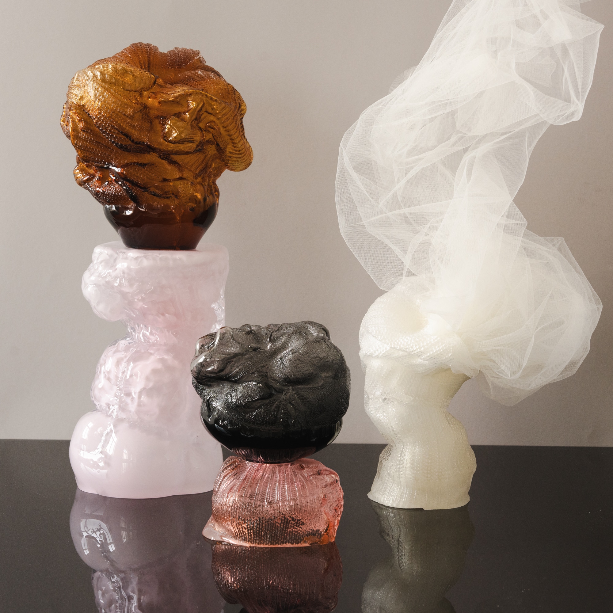 Stacked glass forms on a glossy black surface: amber and smoky grey tops with knitted textures on milky pink and rose bases, and a white knitted form with sheer tulle billowing up from its top.