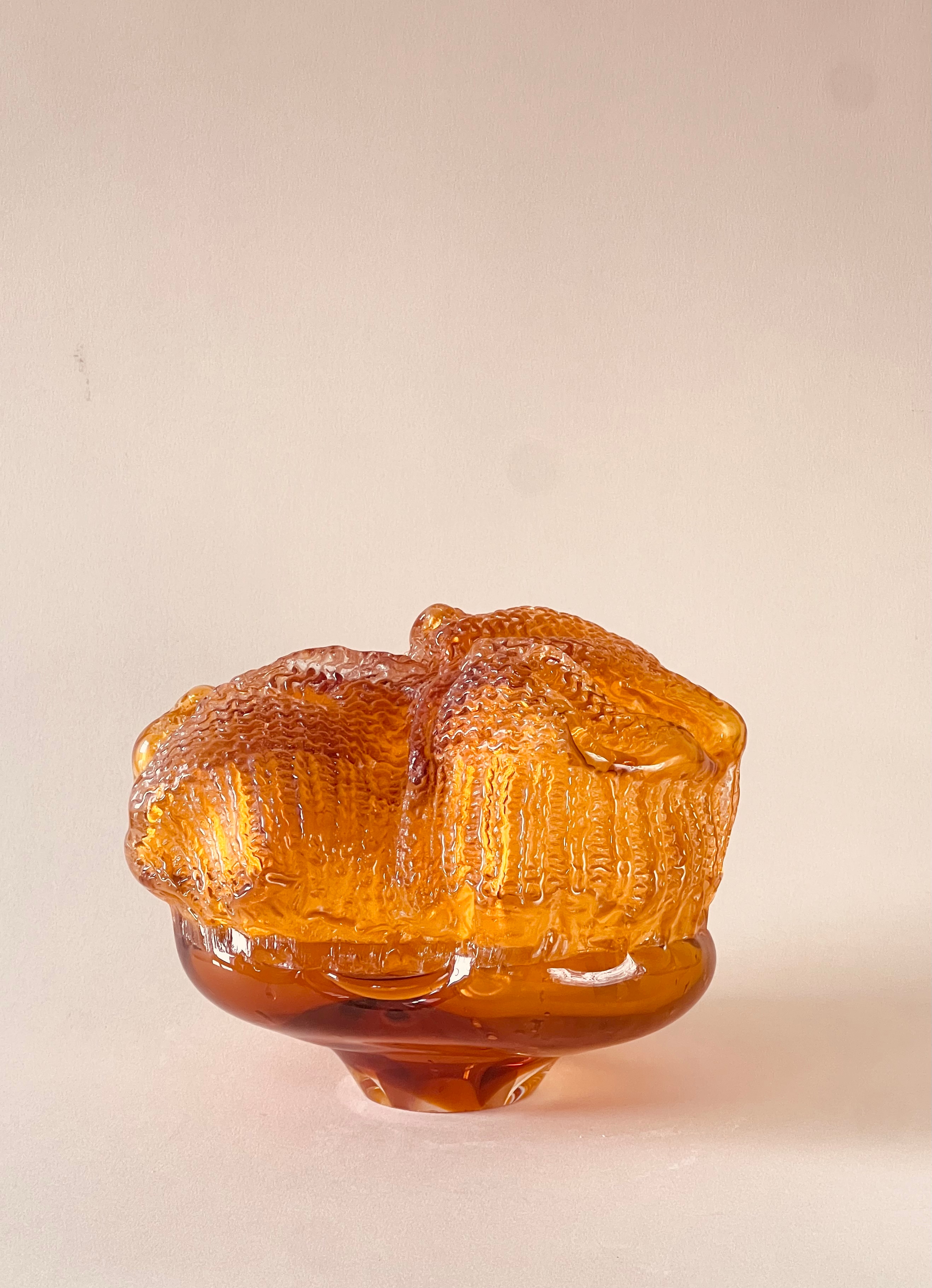 The sculpture Inner monologue: a translucent amber glass form with a knitted texture, folded and gathered over a darker amber base, against a pale pink background.