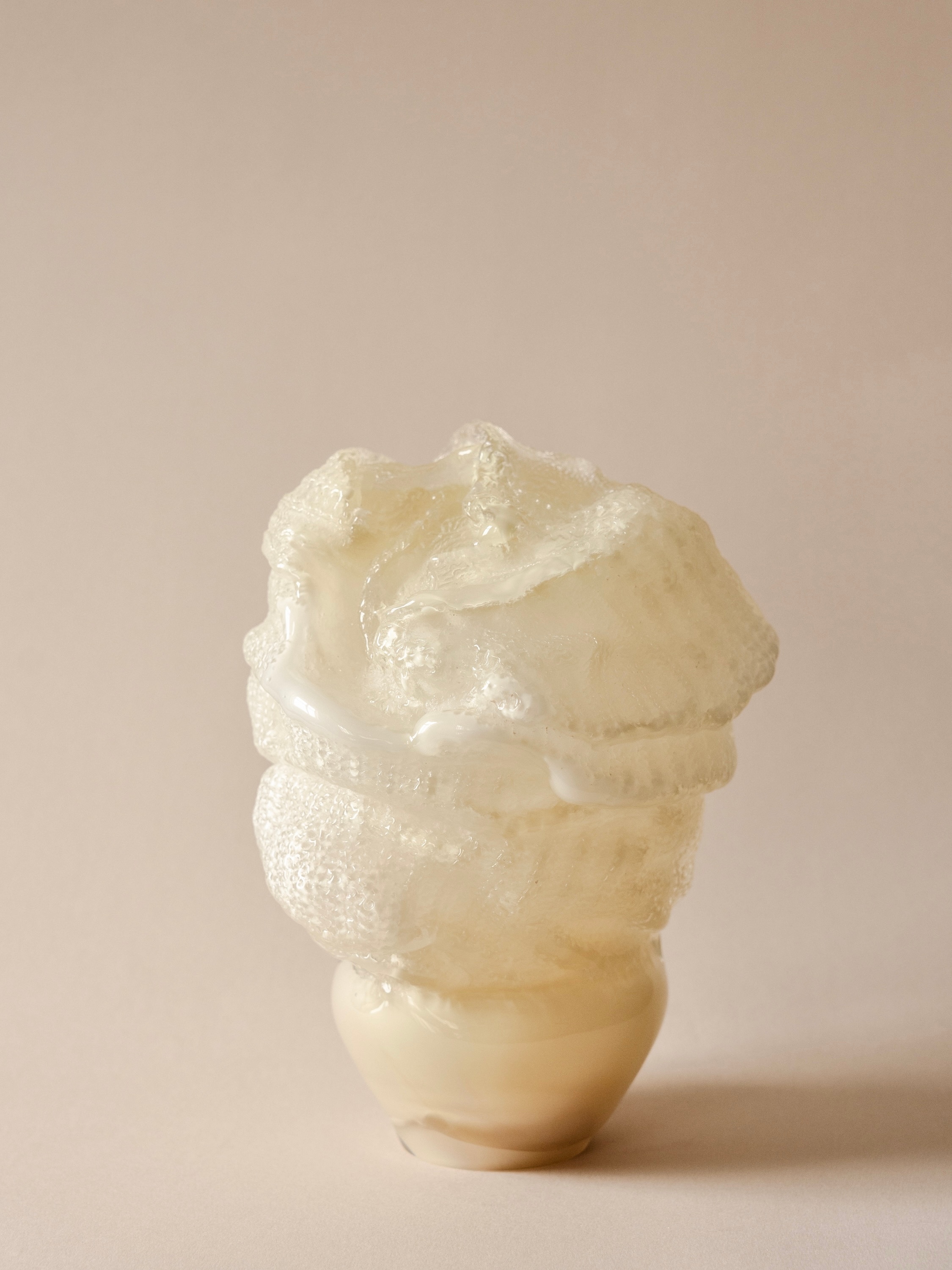 The sculpture Reflections in glass: a milky white and clear glass form with knitted and crocheted textures and a glossy white seam, rising from a cream base, against a pale pink background.