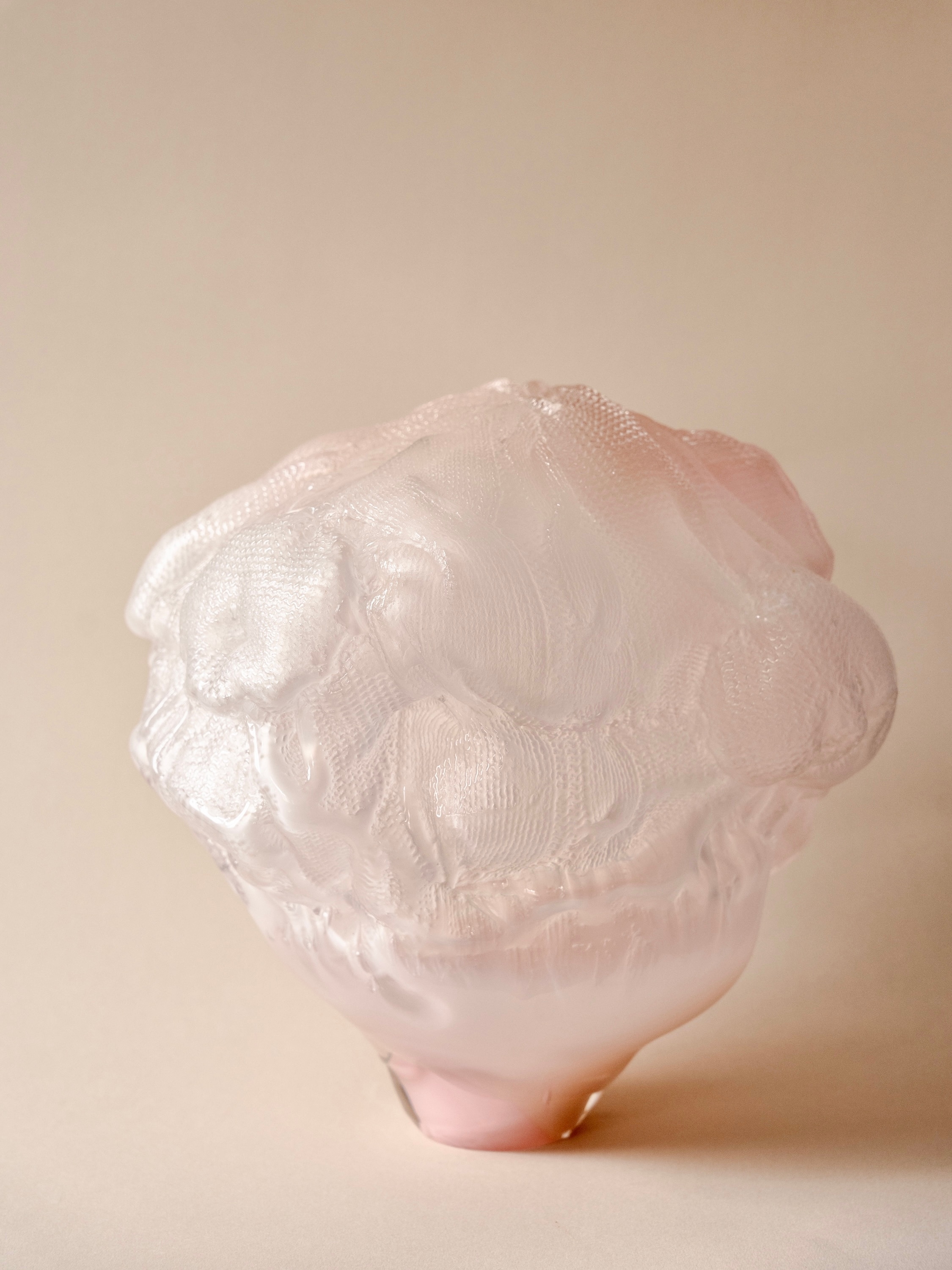 The sculpture Exterior facade: a pale pink and white glass form with knitted and lace-like textures, swelling over a narrow pink base, against a warm beige background.