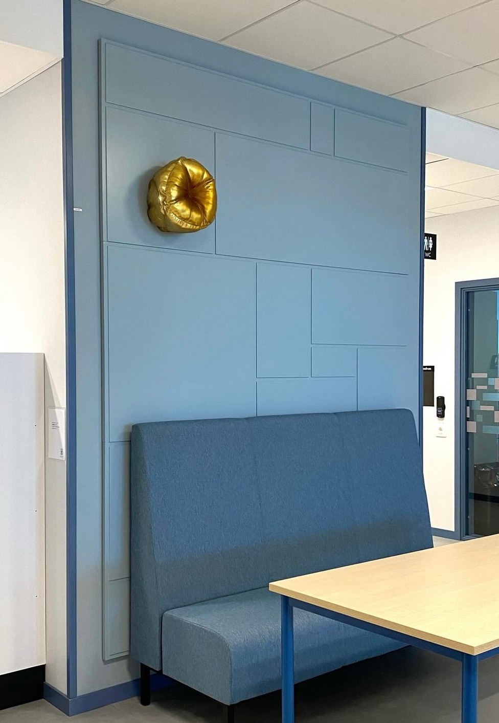 A glossy gold, cushion-like piece with puffed folds gathered to a centre, mounted high on a blue panelled wall above a blue upholstered bench and a light wooden table.