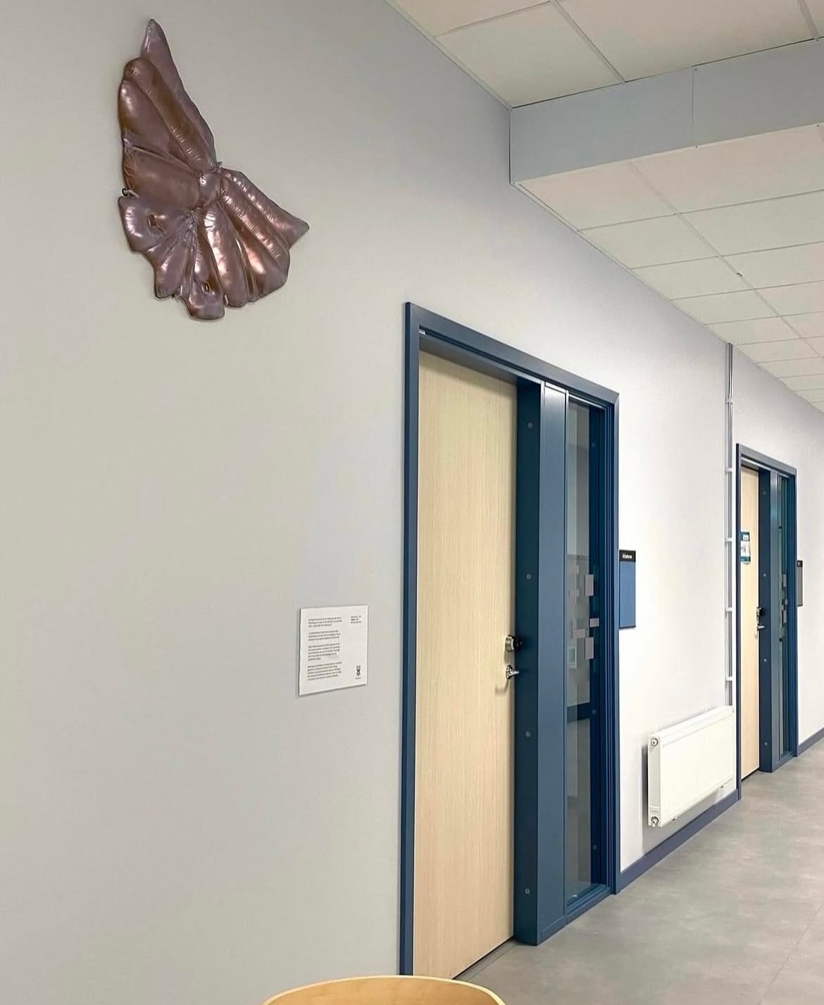A bronze-coloured glass wall relief shaped like spread wings, with puffed, stitched segments, hung high on a pale corridor wall above a text plaque, beside doors with blue frames.