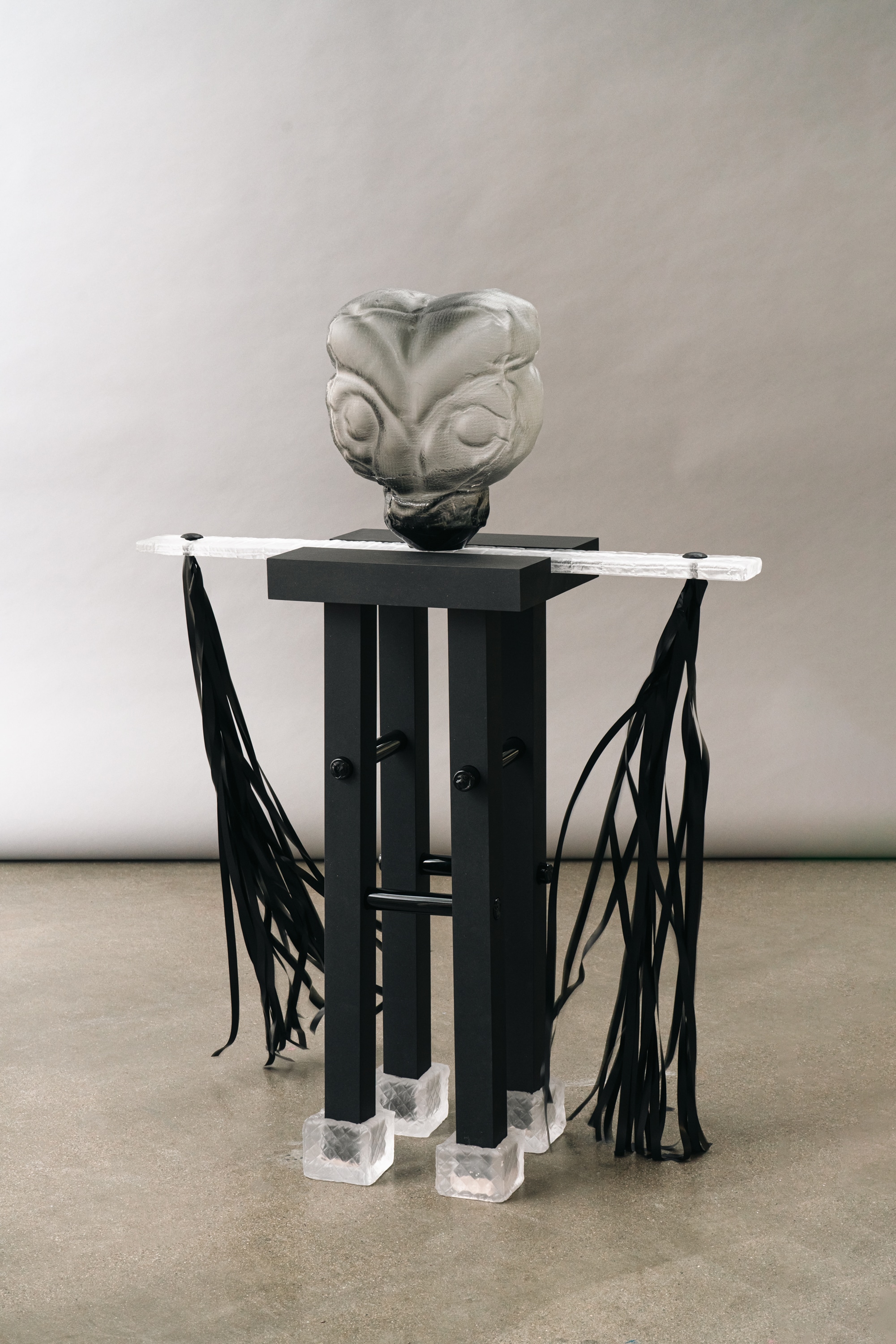 A smoky grey glass head with closed, swollen eyes and a textile-imprinted surface on a matte black stand with frosted glass feet and a clear glass bar hung with black ribbons.
