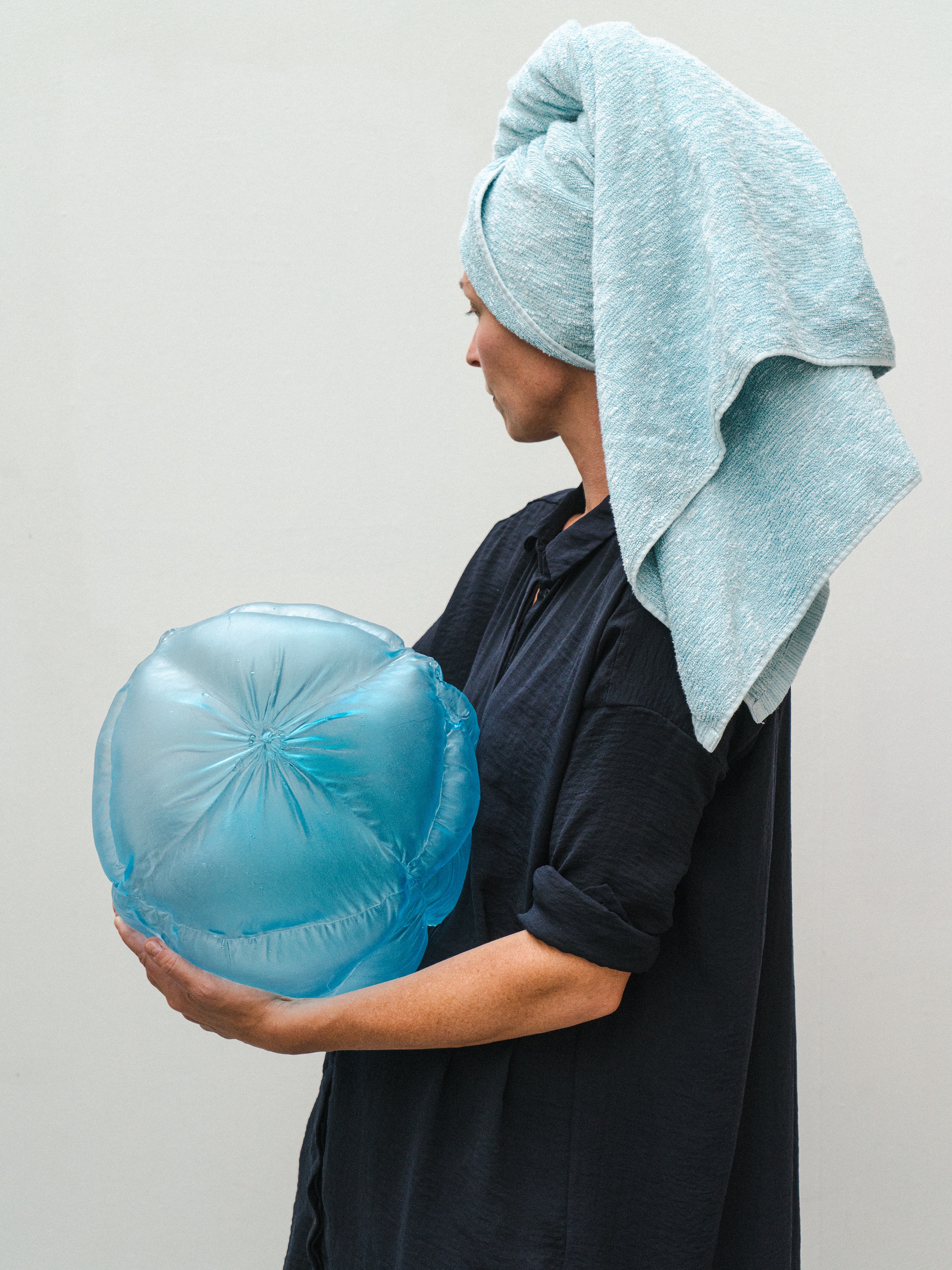 Malin Pierre in profile, a light blue towel wrapped around her head and a navy shirt, holds a round, quilted pale blue glass form against a white wall.