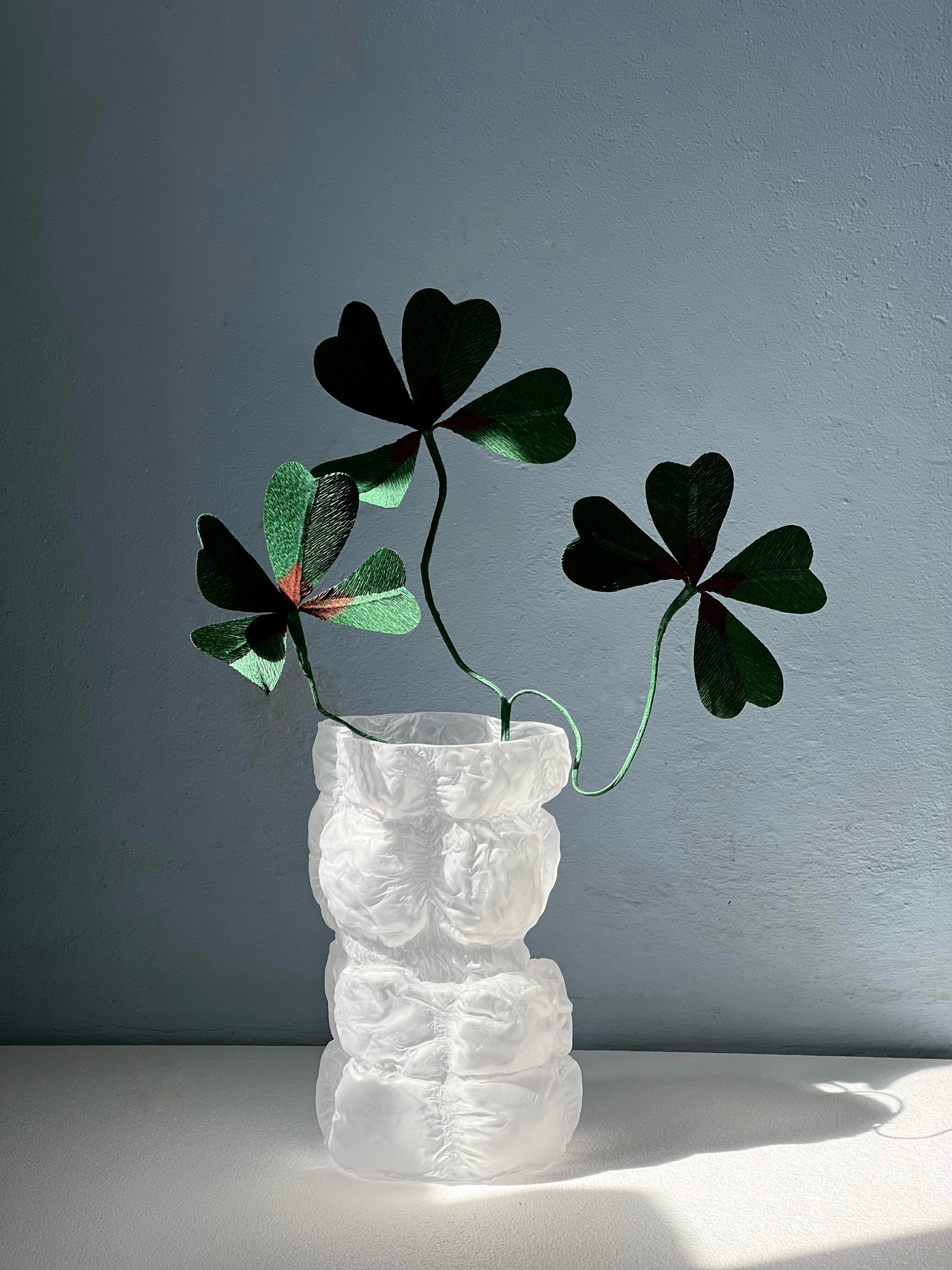 A frosted white vase with a lumpy, stacked form holding green paper flowers against a blue-grey wall.