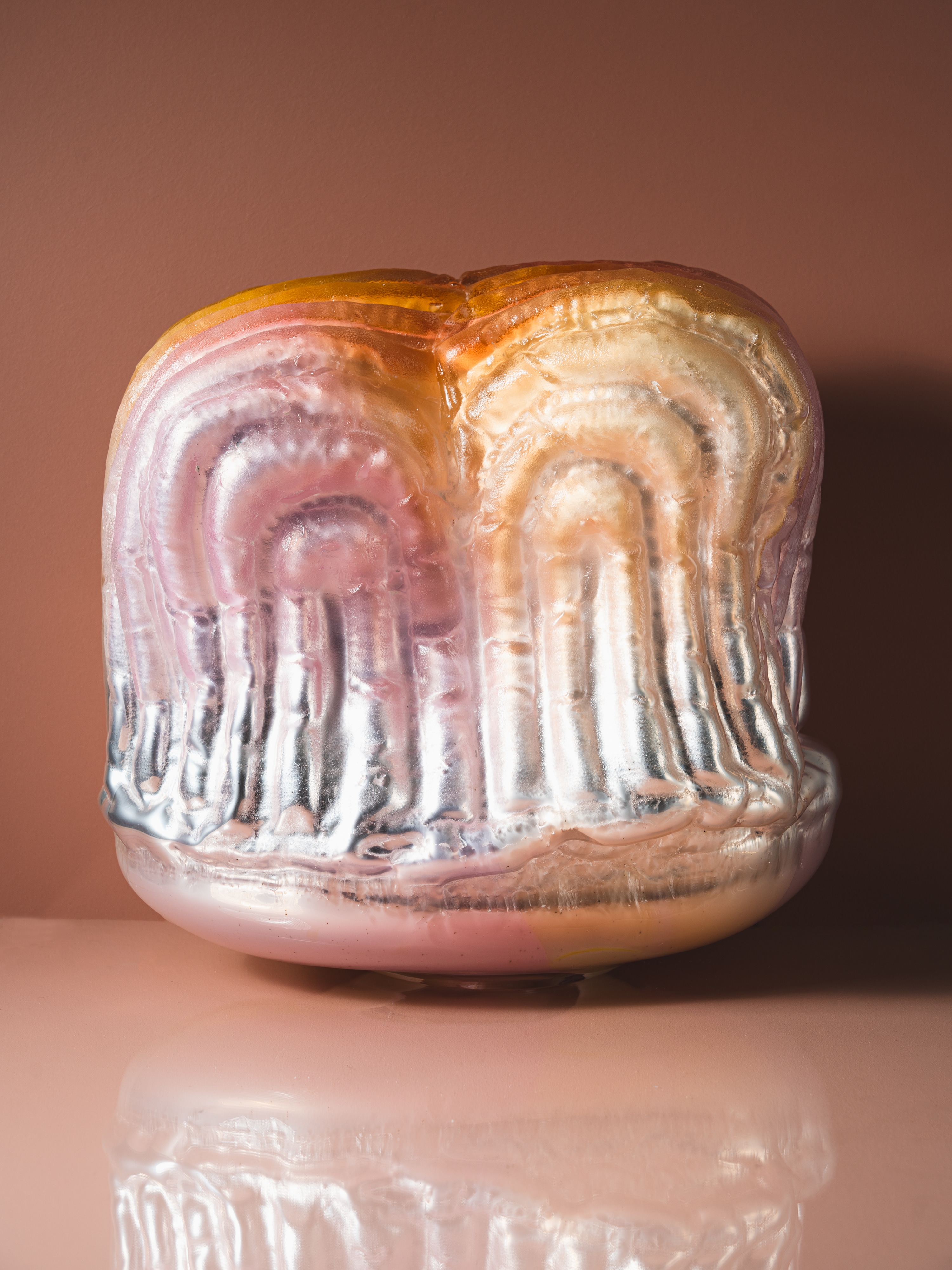 A wide glass form covered in rows of padded arches, pink and amber at the top turning mirror-silver below, on a milky pink base, against a dusky pink wall with its reflection on the glossy surface.