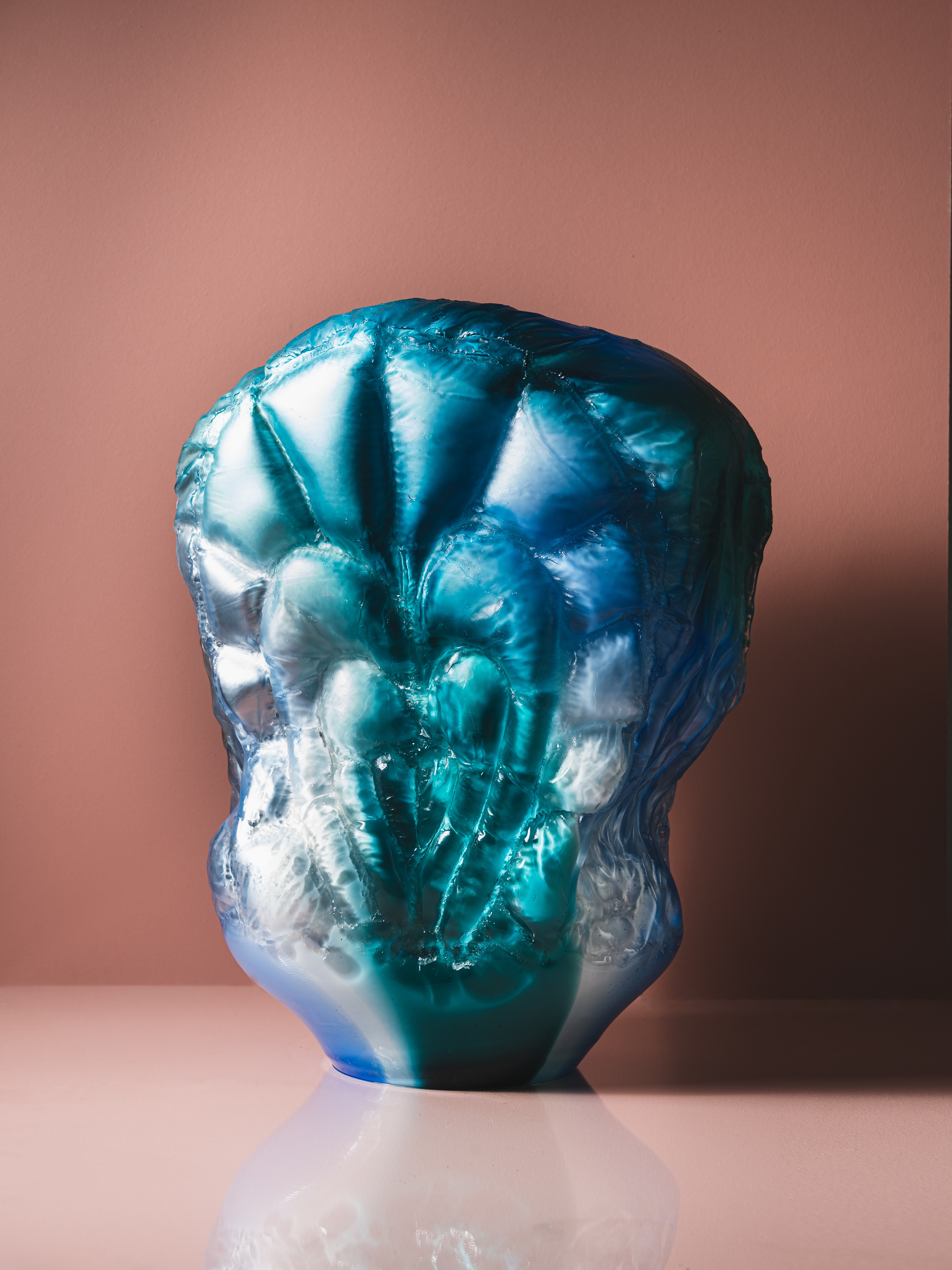 A padded glass form in teal, turquoise and cobalt blue, its puffed, stitched sections fanning out from the centre, on a glossy pink surface against a pink wall.