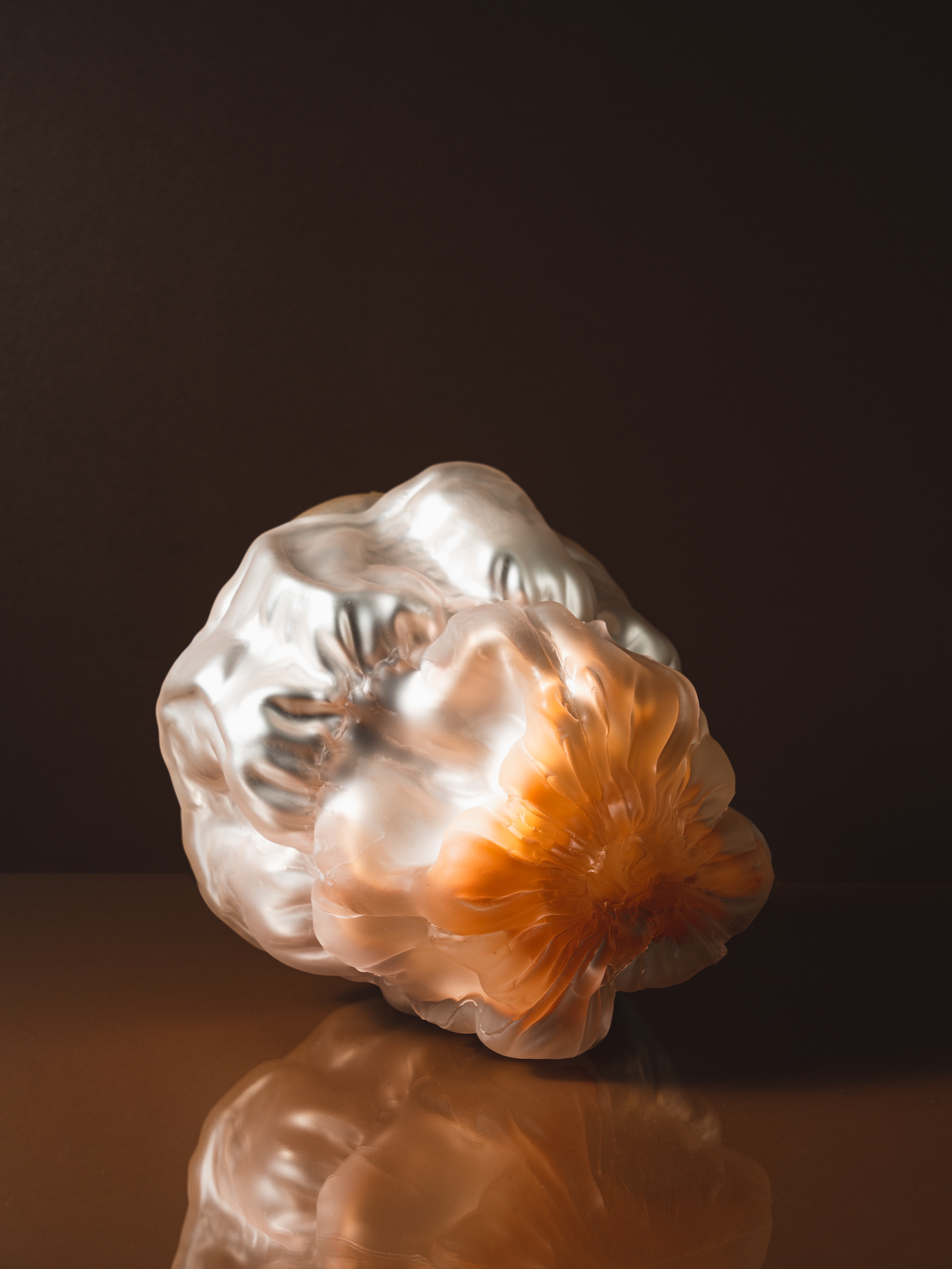A frosted, crumpled glass form lying on its side, clear with a burst of orange at one end, on a glossy brown surface against a dark brown wall.