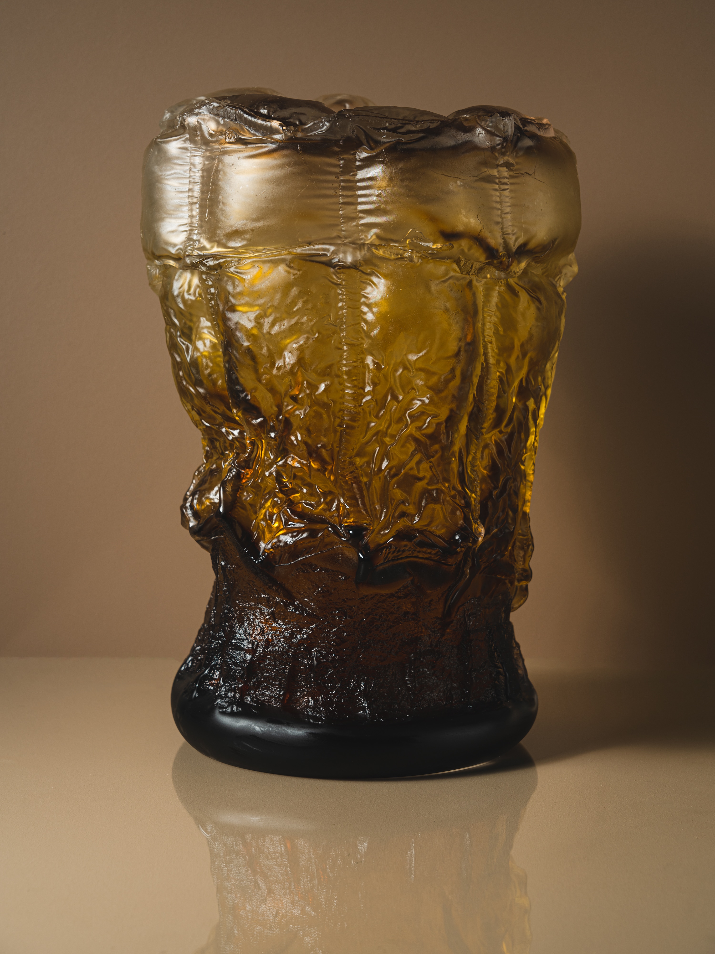 A tall glass vessel with quilted, crinkled walls, pale amber at the padded rim deepening to dark brown at the foot, against a warm beige wall.