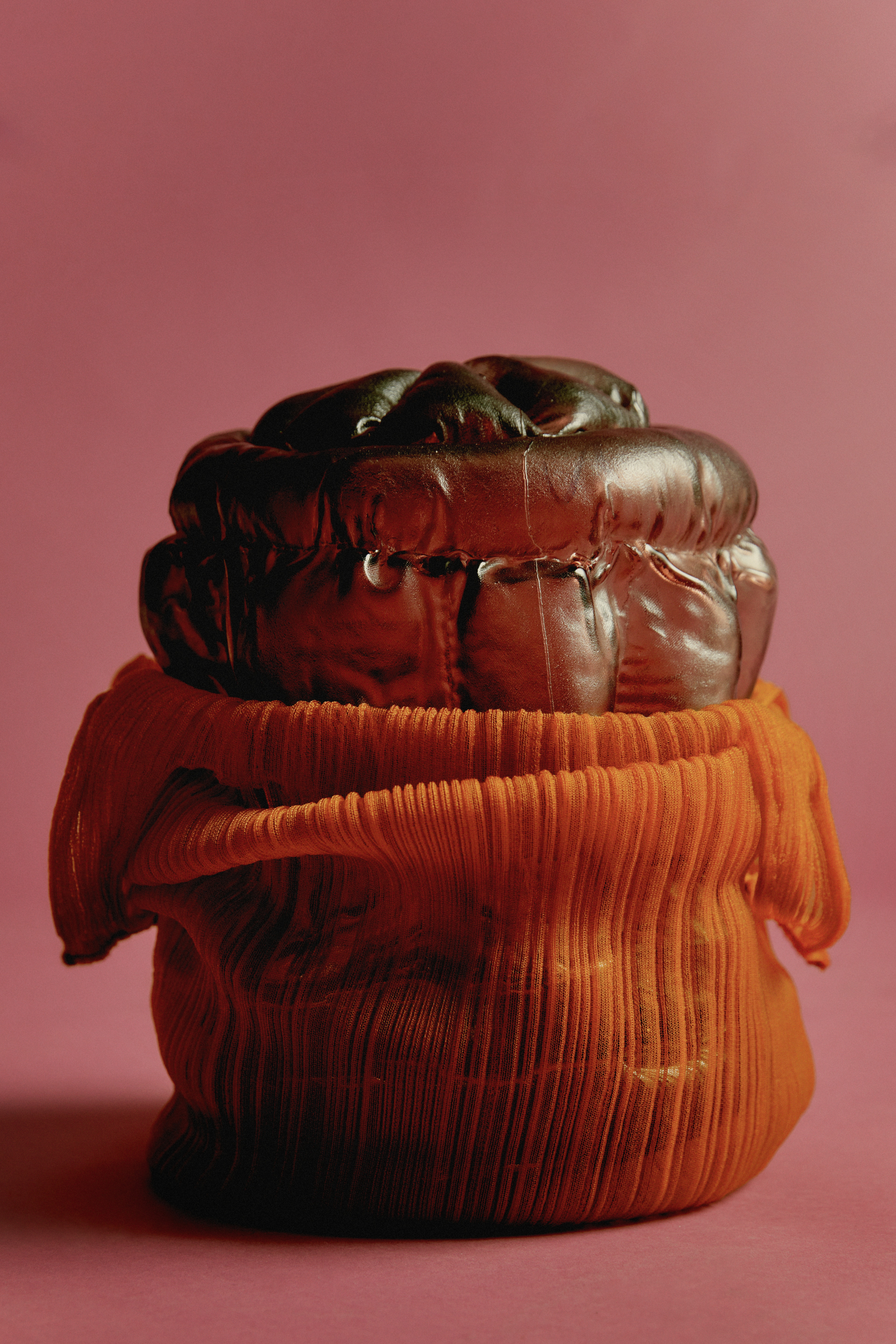 A glossy dark bronze glass form with plump, quilted sections, sitting in a gathered sleeve of finely pleated orange fabric, against a dusky rose backdrop.