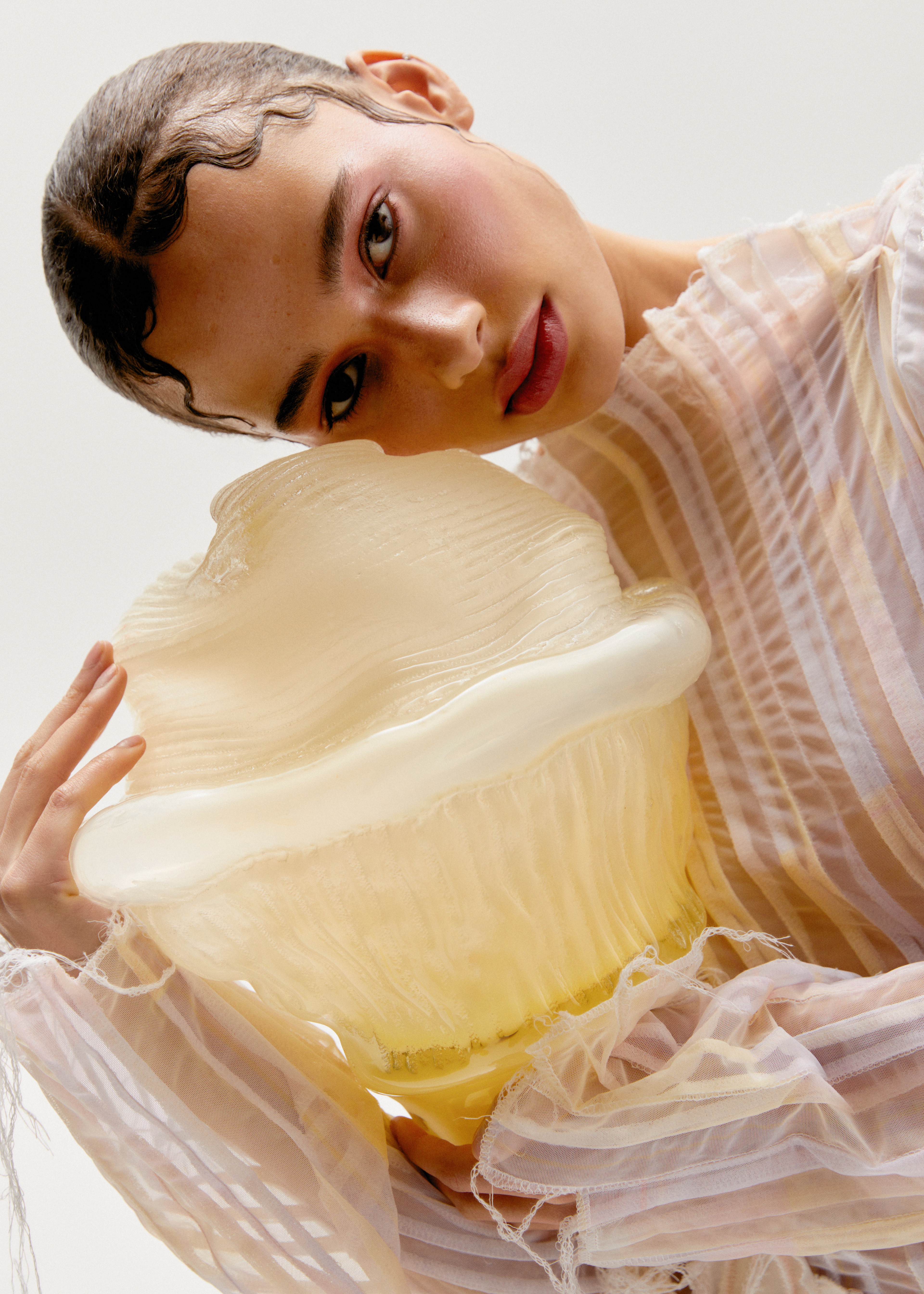 A person in a sheer pink and white striped blouse rests their head on a pale yellow glass form with rippled, ribbed walls and a milky white rim, against a white backdrop.