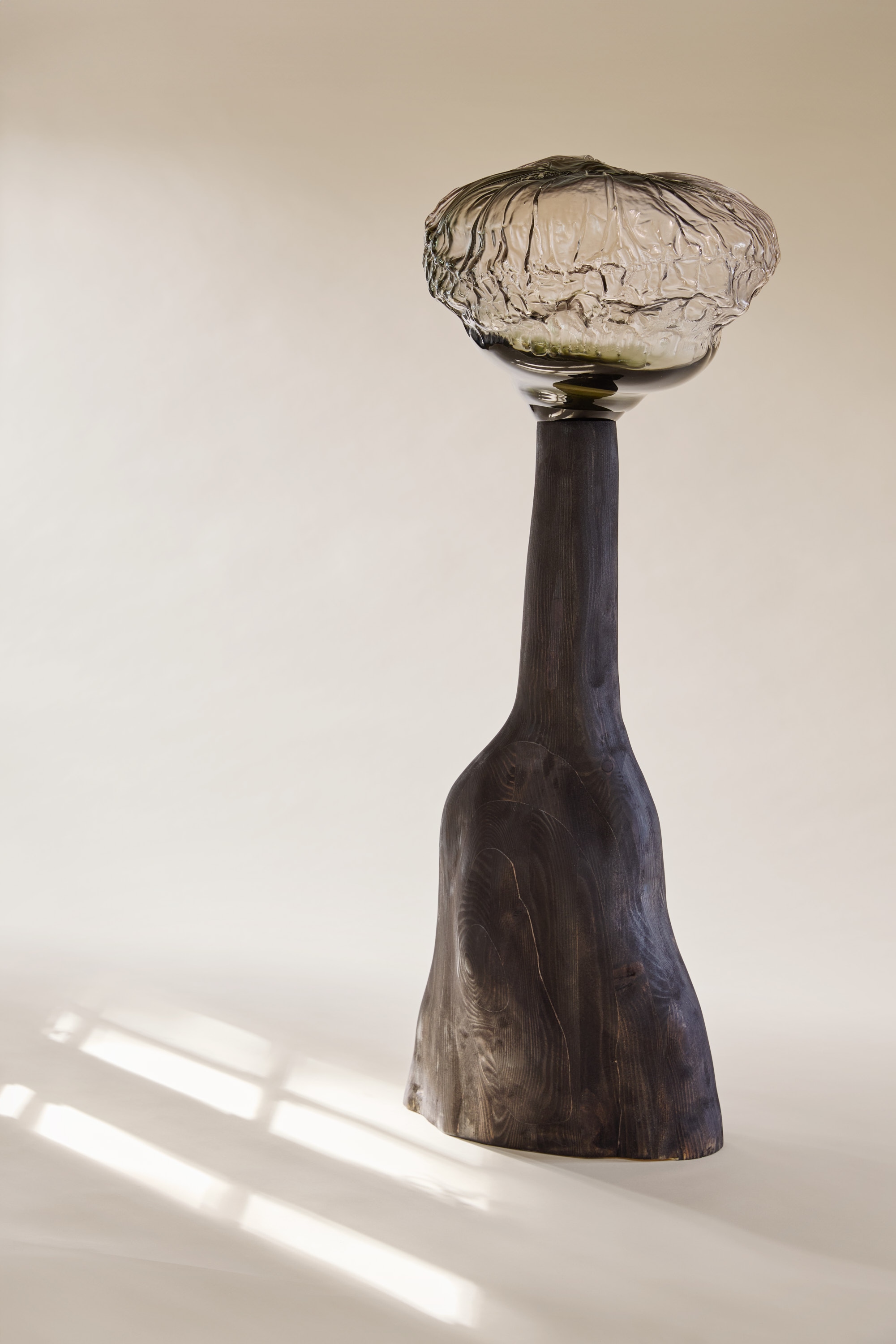 A wide smoky-grey glass top with a crumpled, crinkled surface and glossy underside on a tall charcoal-black turned wooden stem flaring at the foot, with window light on the cream floor.
