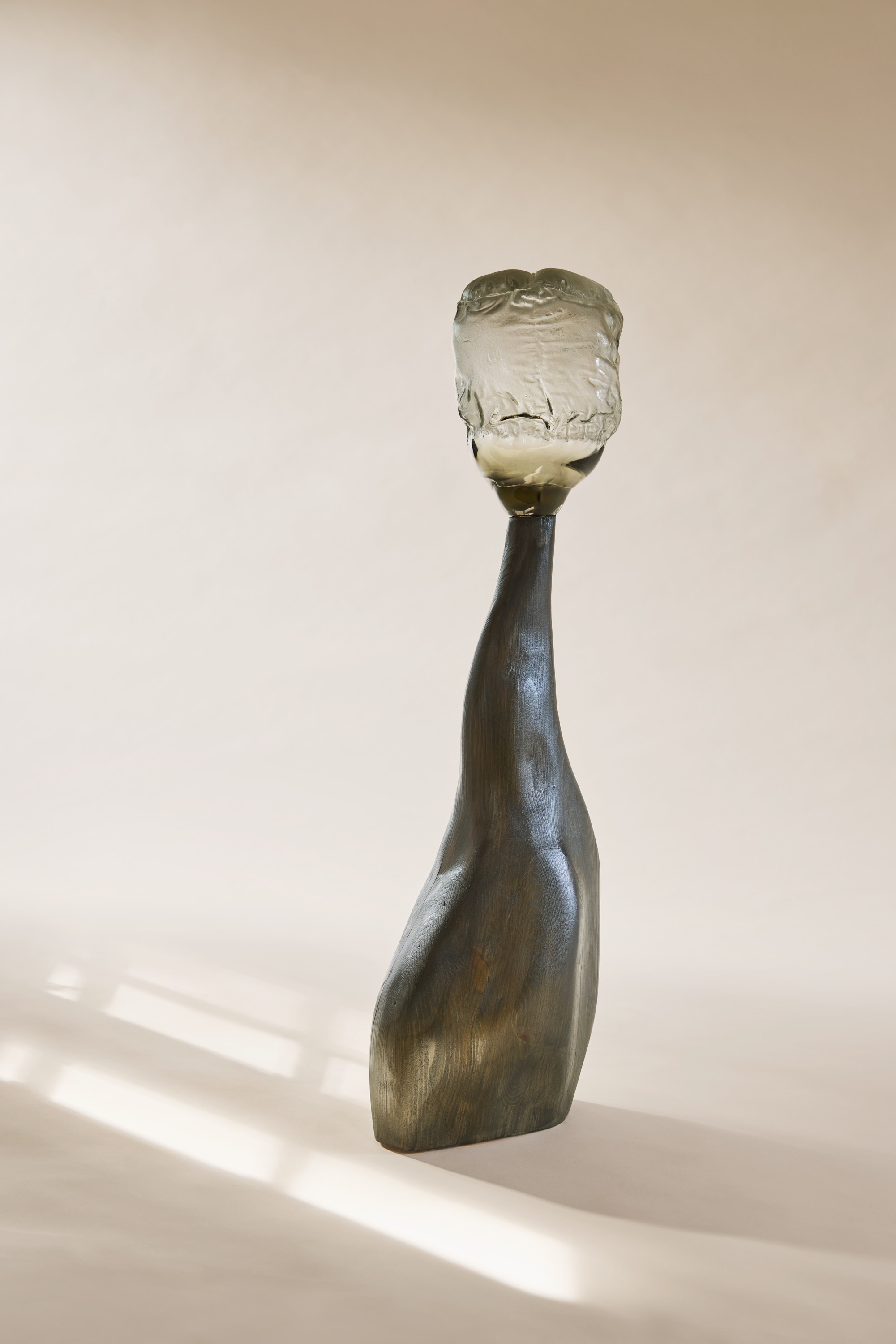 An upright, bud-like glass top, clear with a smoky green-grey tint and a crumpled surface, on a slender leaning wooden stem stained grey-green, against a cream backdrop in window light.