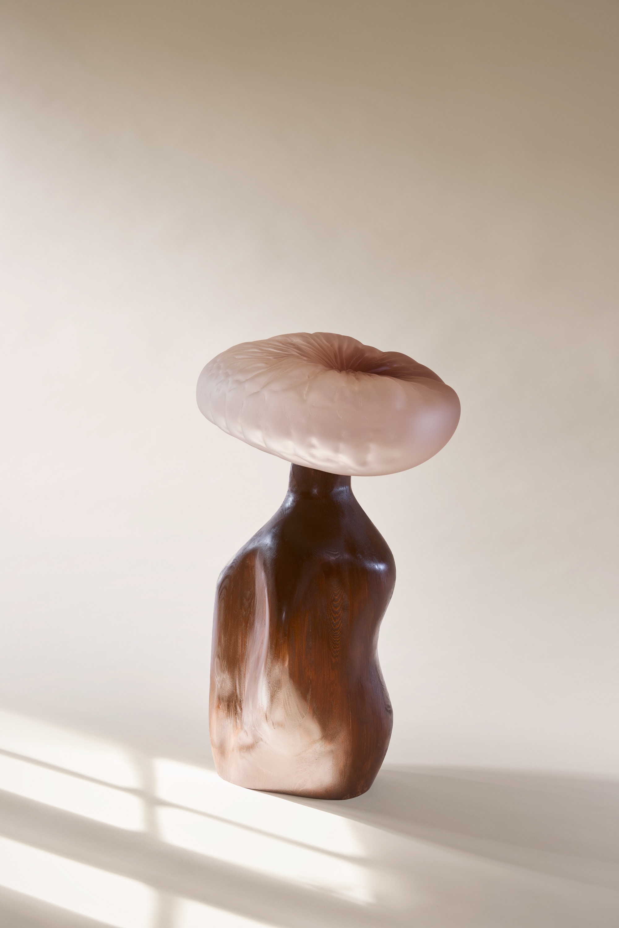 A tilted, cushion-like frosted blush-pink glass cap with ribs radiating from a central dimple, on a chunky turned red-brown wooden stem against a cream backdrop in window light.