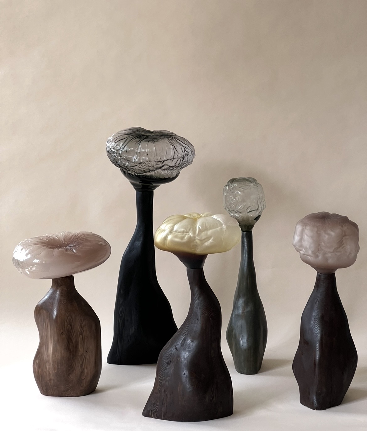 Five sculptures grouped on a pale pink paper backdrop: crumpled or cushion-like glass tops in blush pink, smoky grey, pale yellow, clear grey and mauve on dark turned wooden stems of varied heights.