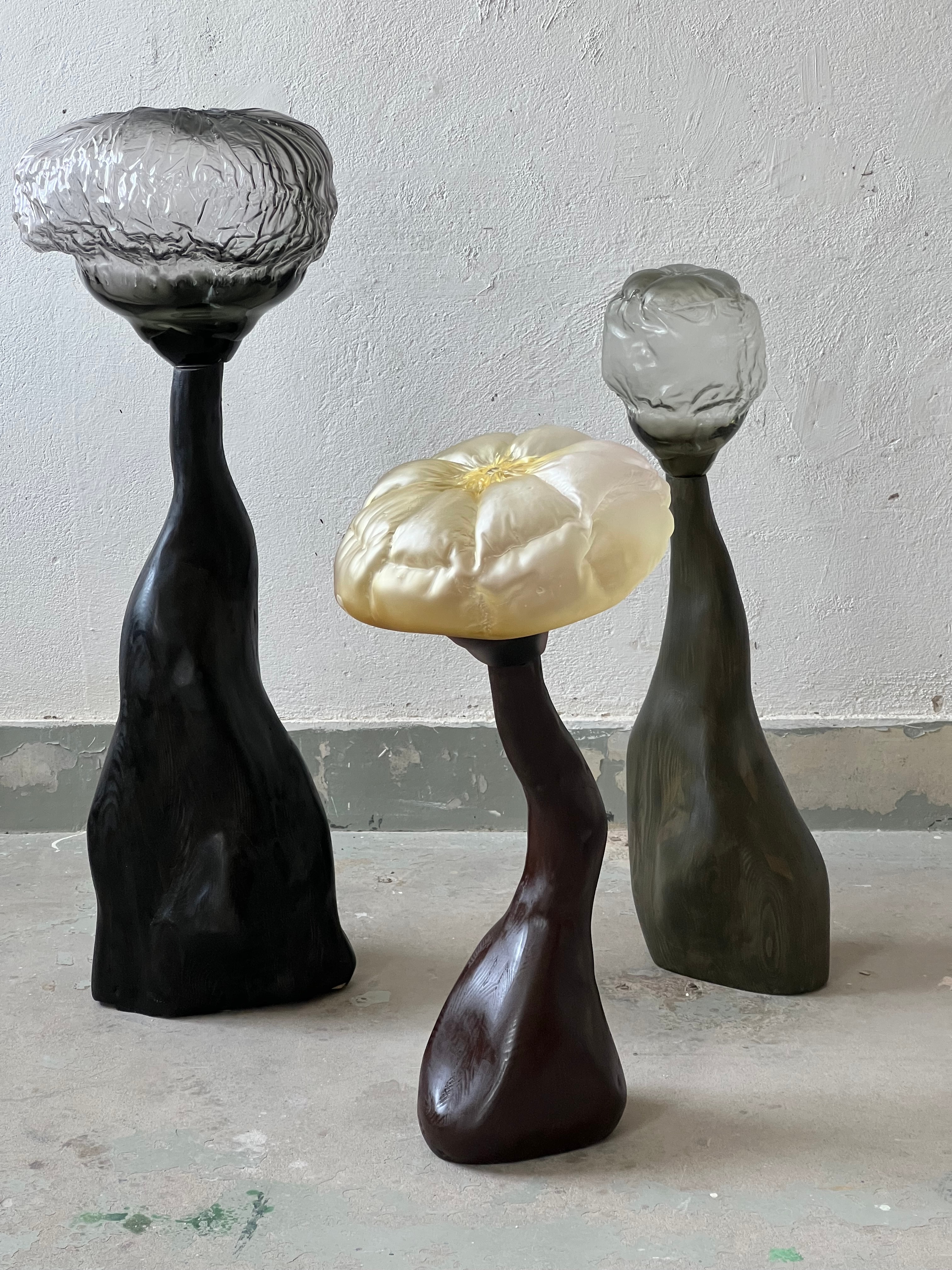 Three sculptures by a rough whitewashed wall on a concrete floor: a pale yellow pillow-like glass top on a dark brown stem between crumpled smoky-grey tops on black and grey-green turned wooden stems.