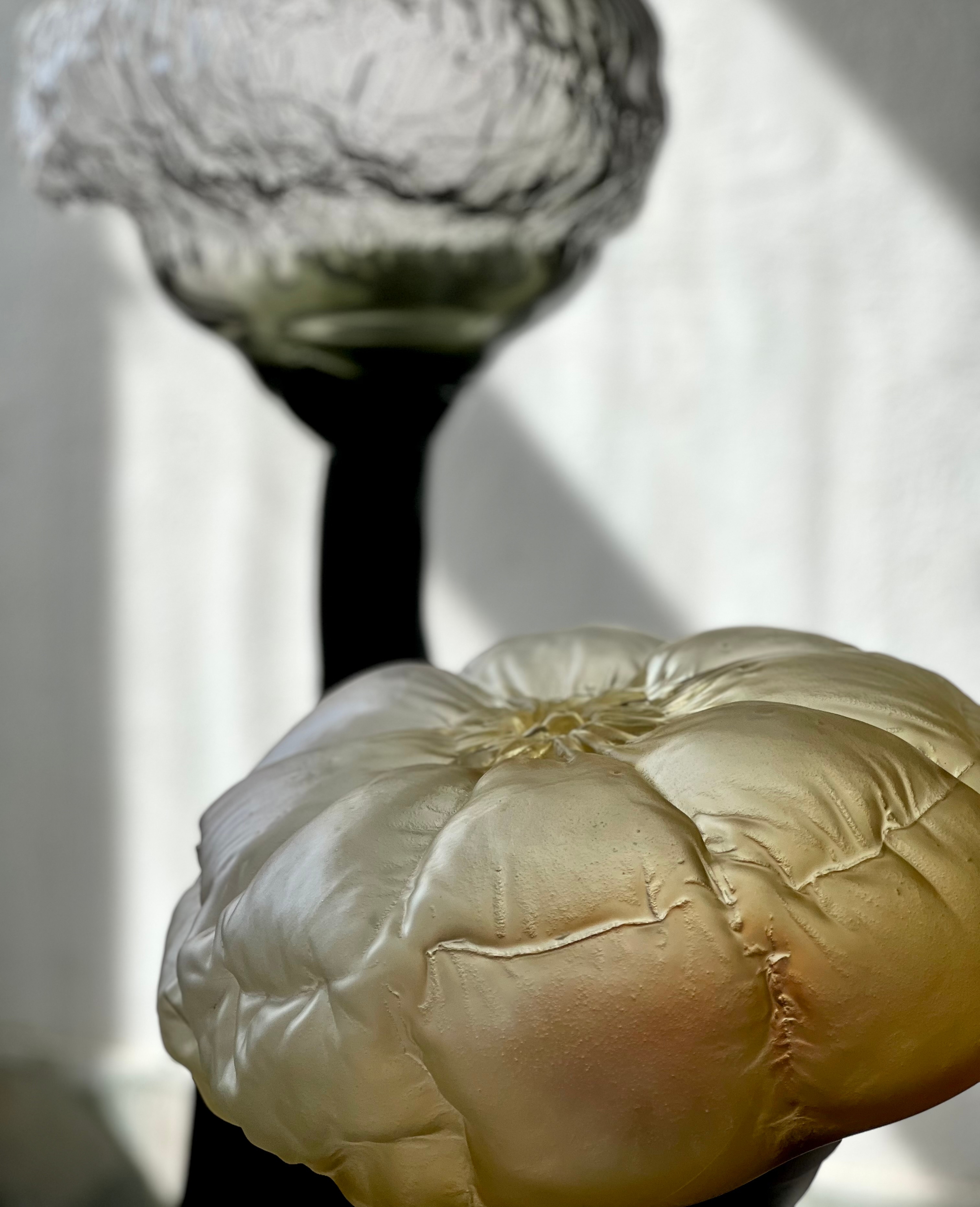 Close-up of a satin pale yellow glass top with plump, pillow-like segments and creased seams; behind it, out of focus, a crumpled smoky-grey top on a dark stem in sunlight.