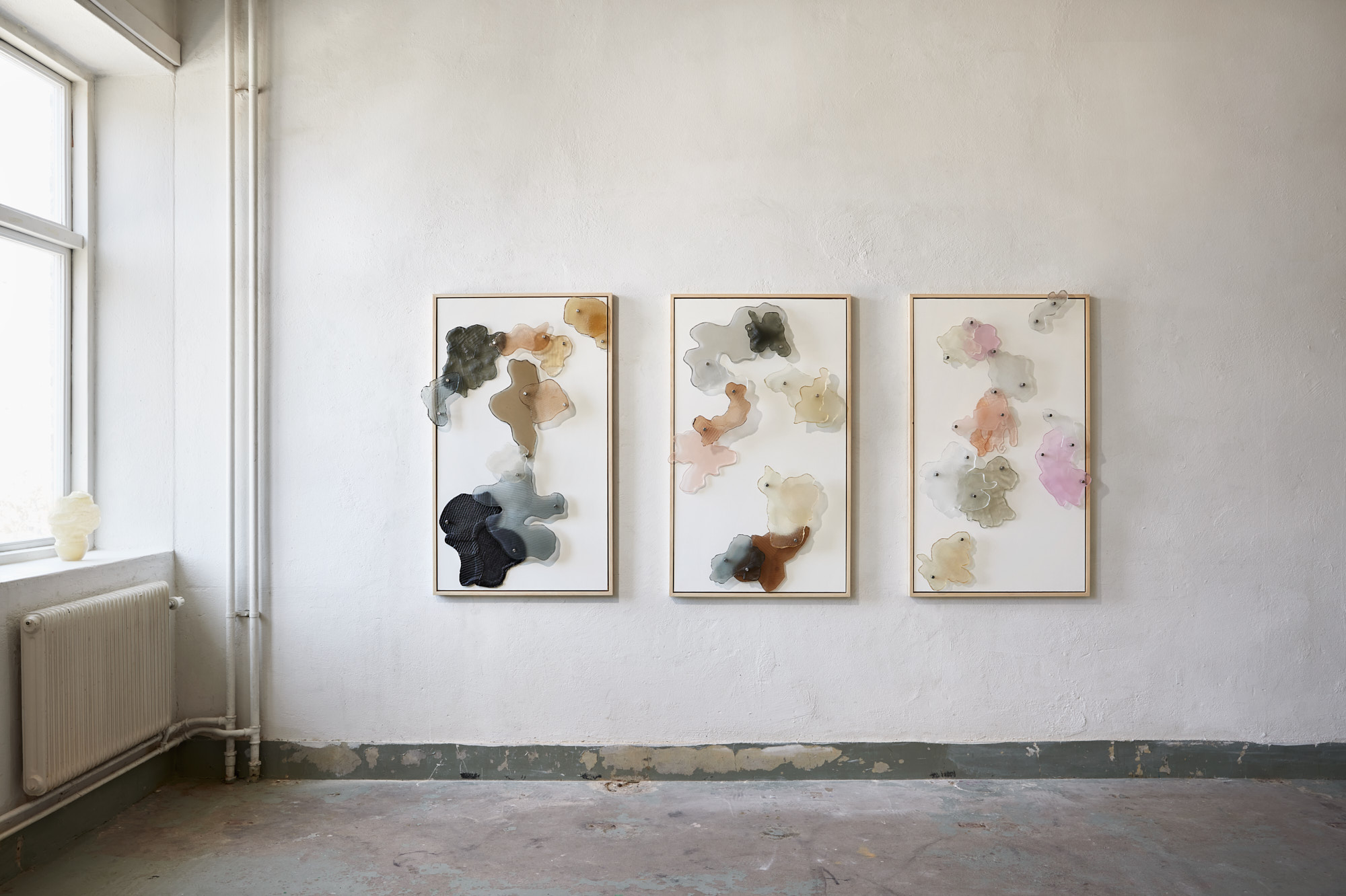 Three tall light-wood frames on a whitewashed gallery wall, each scattered with translucent glass shapes in black, grey, amber, peach, pink and cream, with a window at left.