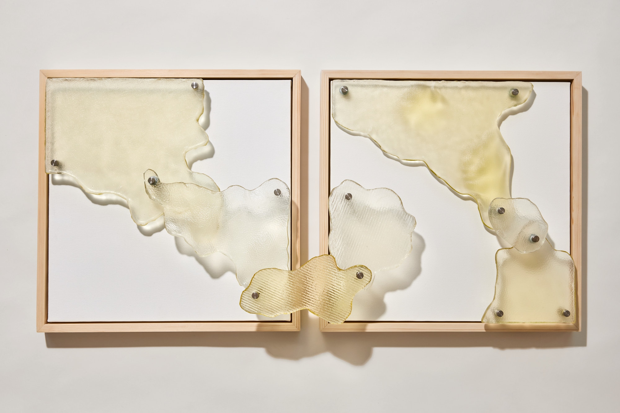 Two square light-wood frames on a white wall, with pale yellow and clear glass shapes, some ribbed or textured, pinned on steel spacers across white canvas and over the frame edges.