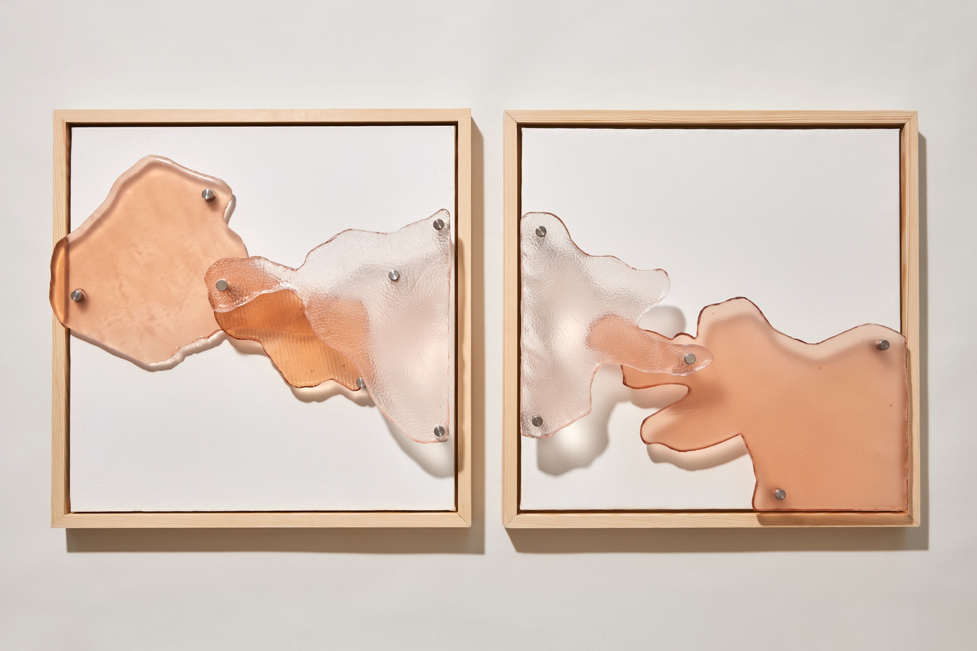 Two square light-wood frames on a white wall, with peach and clear textured glass shapes pinned on steel spacers across white canvas, casting soft shadows.