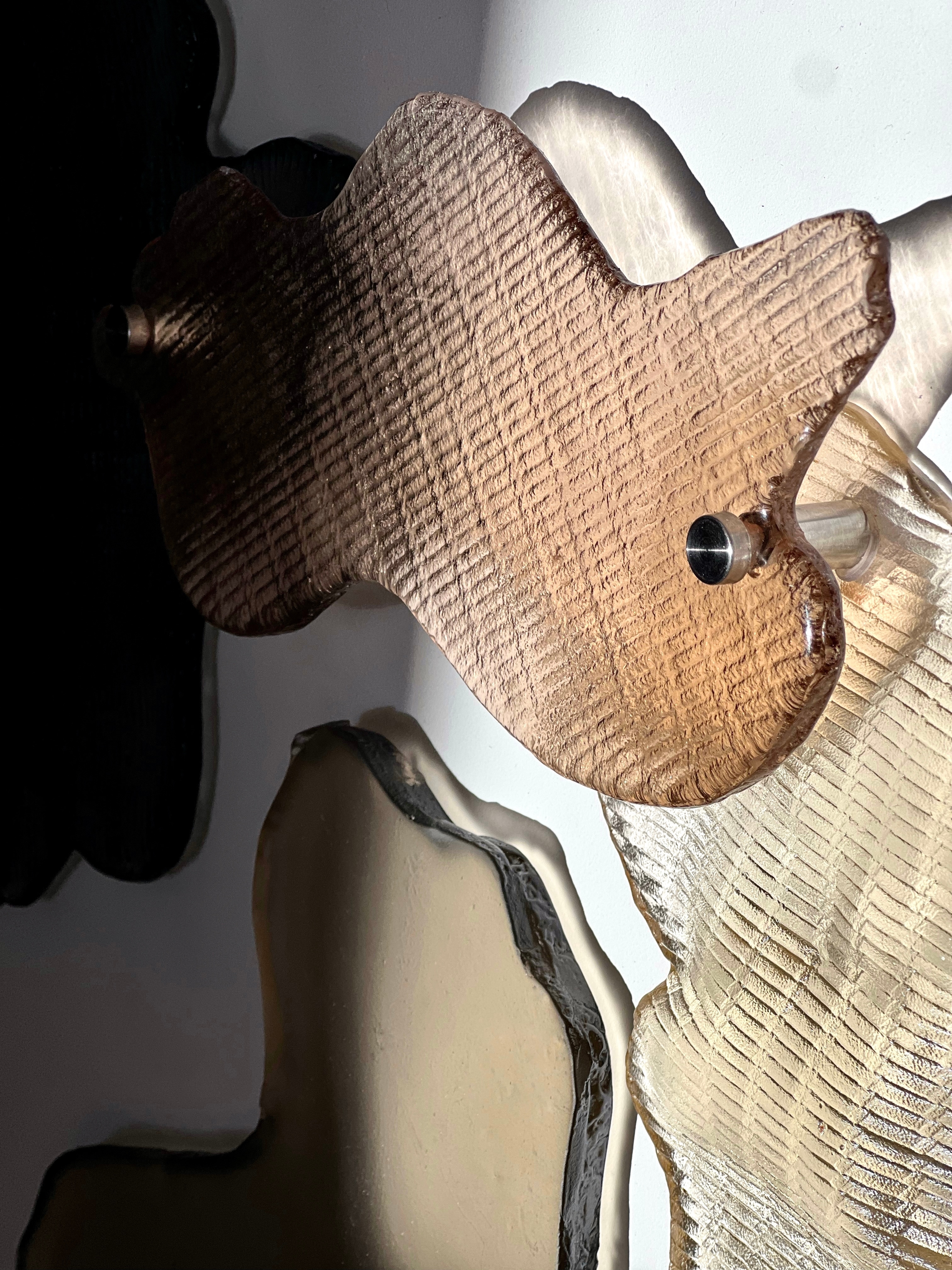 Close-up in hard side light: a smoky brown glass shape with fine ribbed texture on a steel spacer, over a pale gold patterned piece, with a black ribbed piece at left and a smooth grey one below.