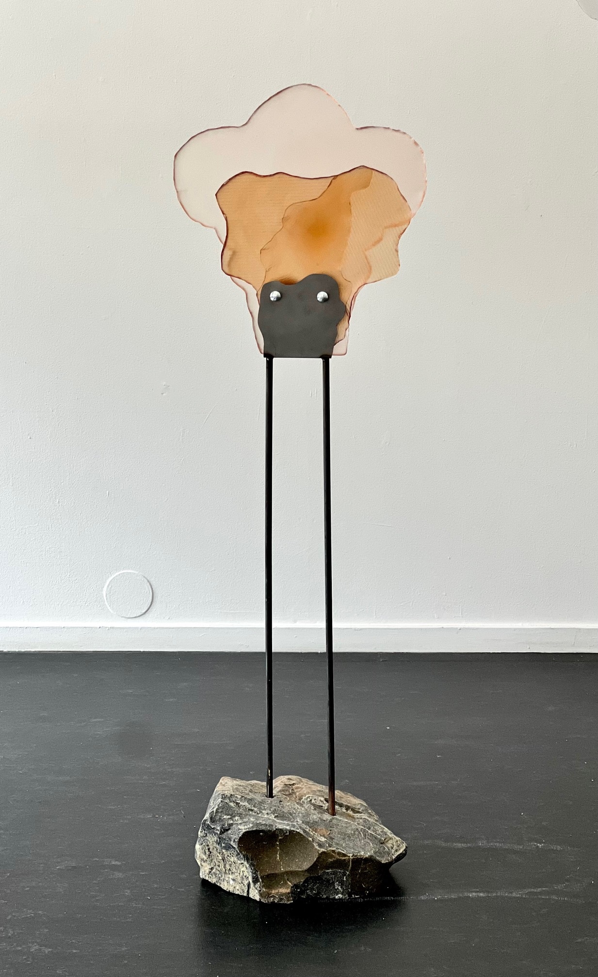 A standing sculpture: layered pale pink and amber glass shapes like a cloud, held by a dark metal plate on two thin black rods rising from a rough grey stone on a dark floor.