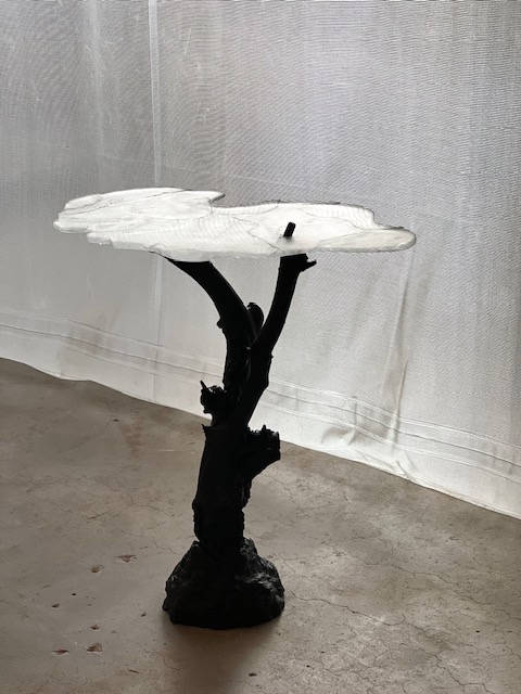 The sculpture Skogsdimma: a frosted white glass top like a drifting cloud on a black, branch-like stand with a rough base, on a concrete floor in front of a sheer white curtain.