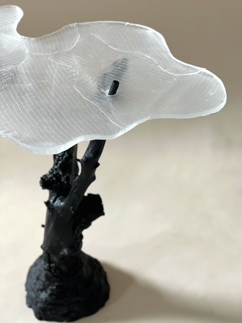 Close-up of Skogsdimma: the frosted glass top, finely ribbed like contour lines, pinned to the knotted black branch-like stand, against a pale beige wall.