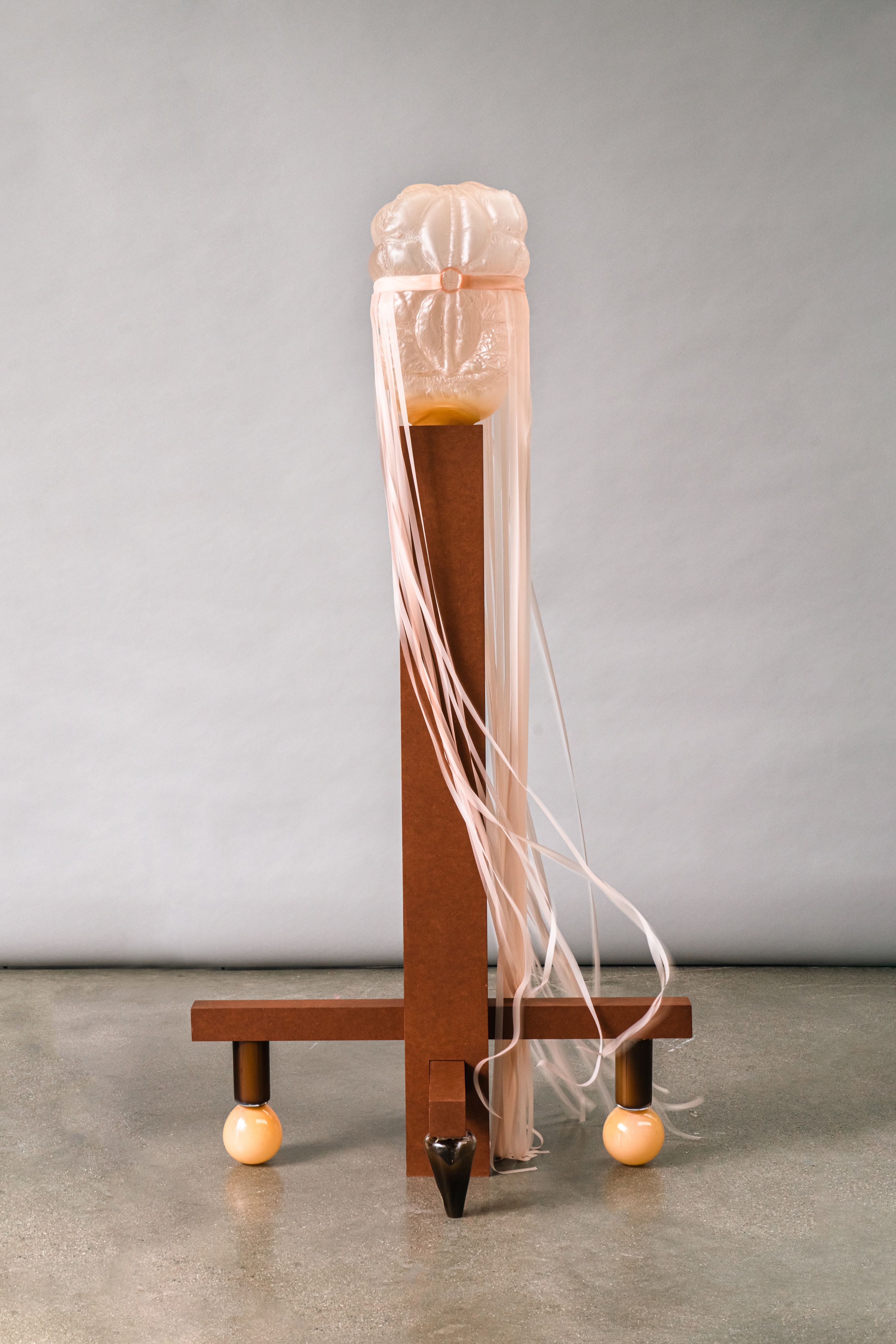 A translucent peach glass form with quilted, puffed panels, belted with a pink ribbon that trails to the floor, on a tall brown wooden stand with glossy peach glass ball feet.