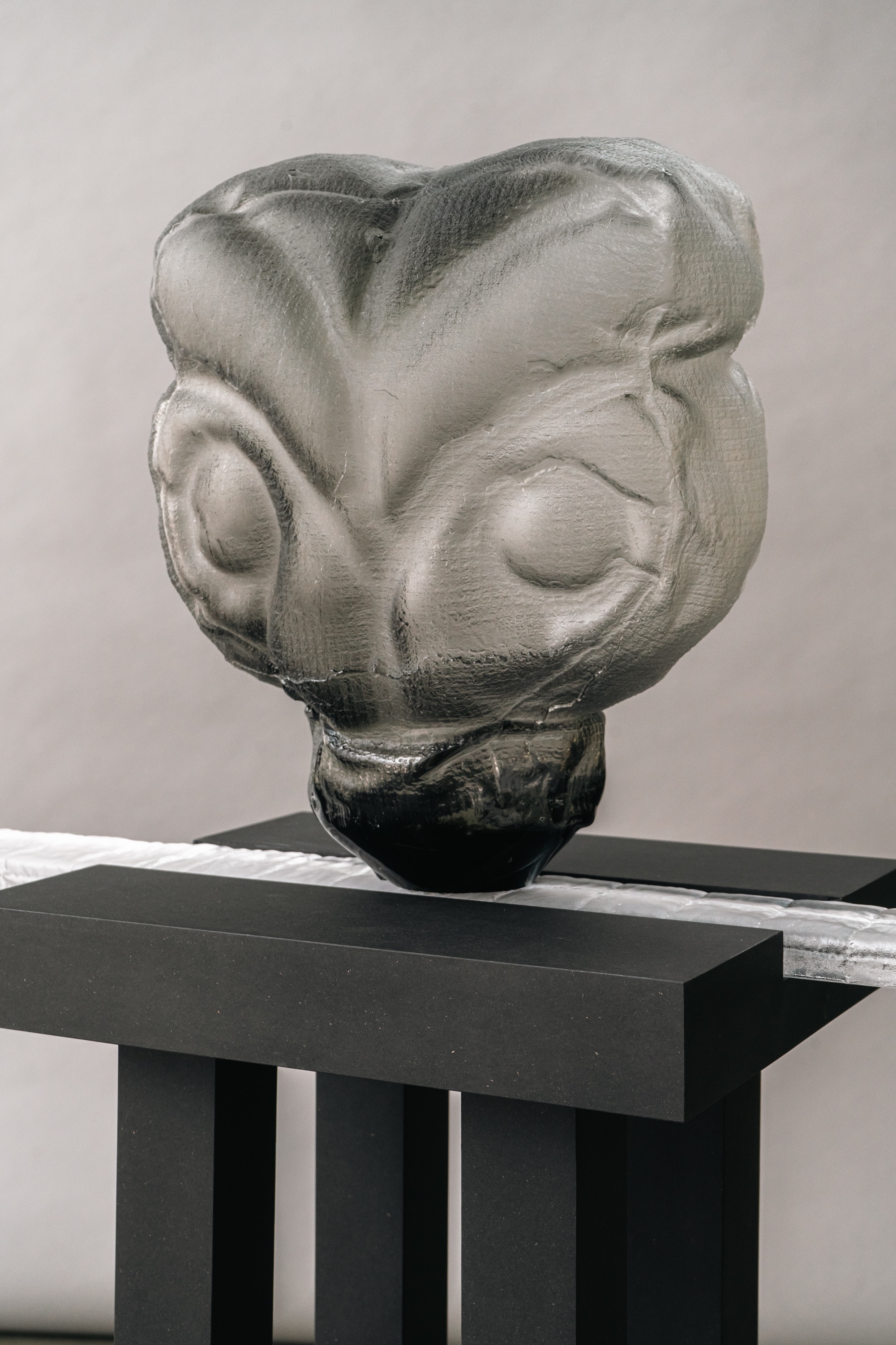 Close-up of a smoky grey glass head with closed, swollen eyes and a fine woven-textile imprint, darkening to black at its base, resting on a matte black stand and a clear glass bar.